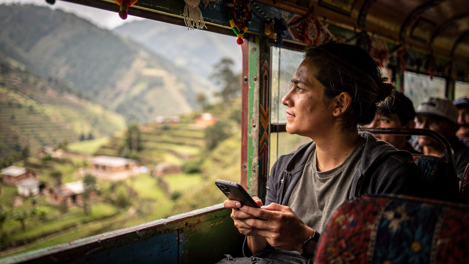 Un viajero consulta su teléfono mientras el autobús local atraviesa el paisaje montañoso del interior de Guatemala, con pueblos pequeños y campos de cultivo en la distancia.