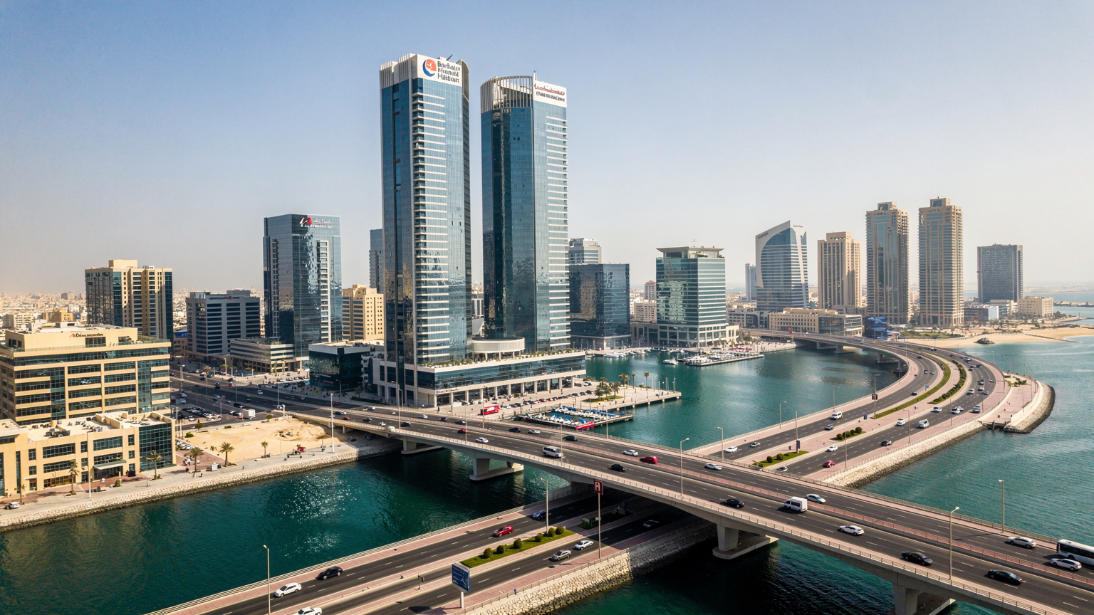 صورة جوية لخليج البحرين التجاري، تظهر فيها ناطحات السحاب الحديثة على الواجهة البحرية، مع حركة مرور خفيفة على الطرق السريعة المجاورة. إضاءة نهارية صافية.