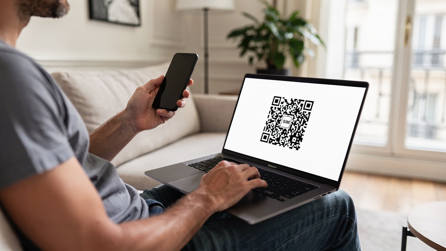 A traveler in a French apartment living room, holding a smartphone with an eSIM QR code displayed on a laptop screen, ready for scanning.