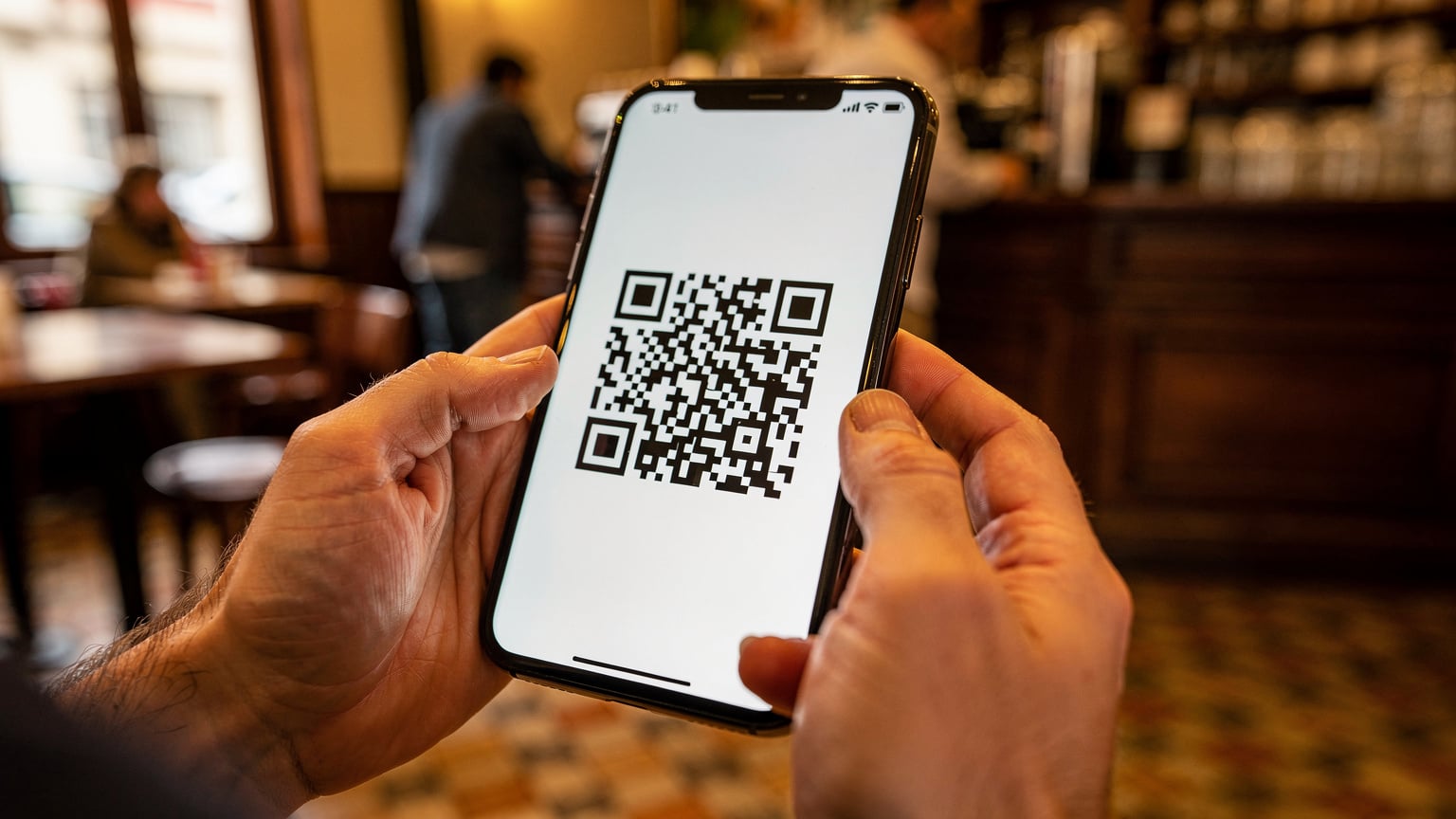 Primer plano de unas manos sosteniendo un smartphone, mostrando un código QR en la pantalla para activar una eSIM, con un fondo ligeramente desenfocado de un café en la Ciudad Vieja de Montevideo.