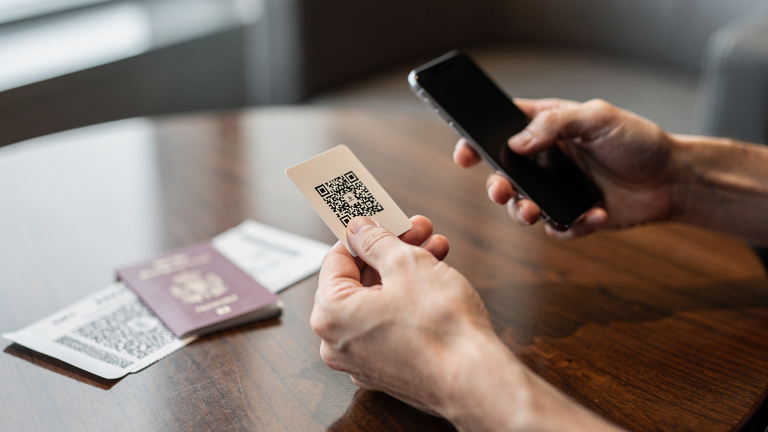 Un primo piano delle mani di una persona che scansiona un codice QR sul telefono per attivare una eSIM, con un passaporto italiano e una carta d'imbarco sfocati sullo sfondo su un tavolo di legno in un salotto d'aeroporto.