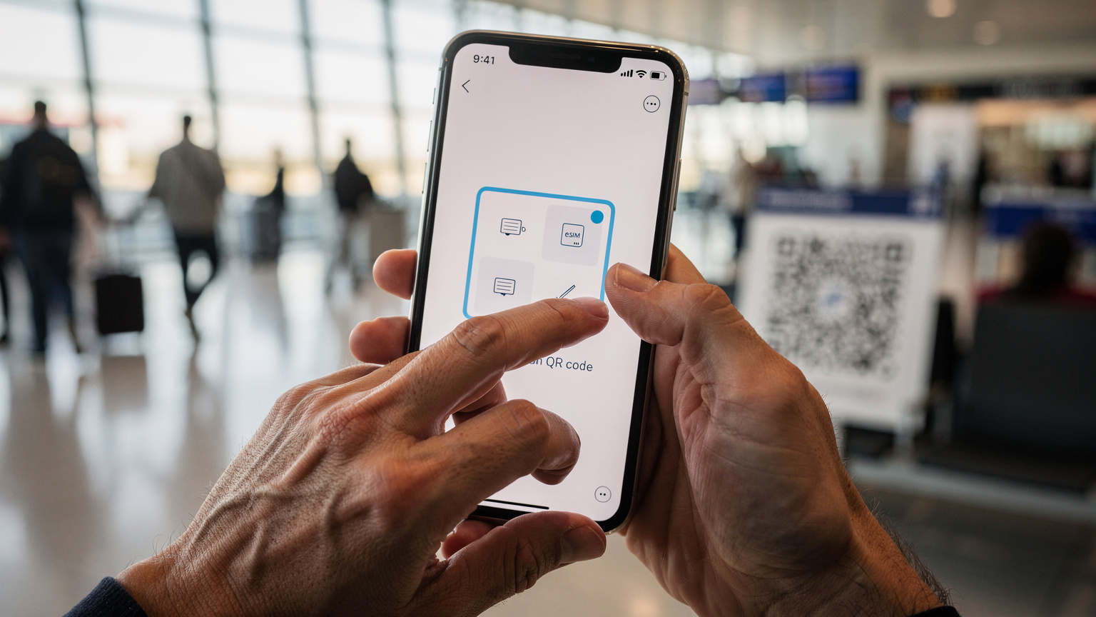 Un gros plan sur les mains d'une personne qui tient un smartphone, affichant l'écran de configuration d'une eSIM avec un QR code à l'arrière-plan flou, dans un terminal d'aéroport bondé avec une lumière diffuse de l'après-midi.