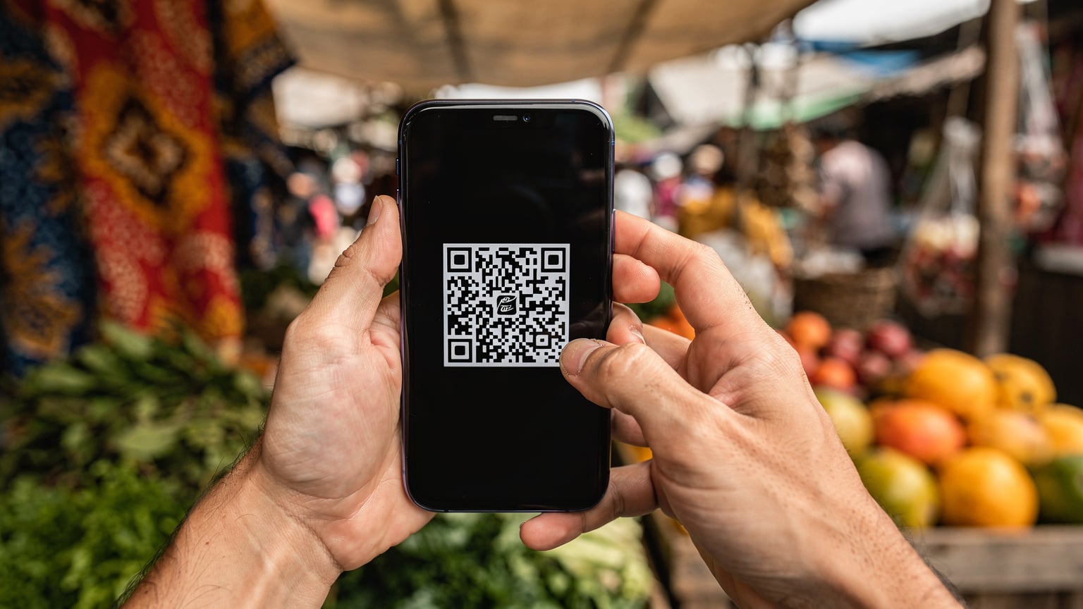 A close-up of a traveler's hands holding a smartphone, fingers hovering over the screen, displaying a Cellesim eSIM activation QR code, with a blurred backdrop of a traditional Indonesian market.