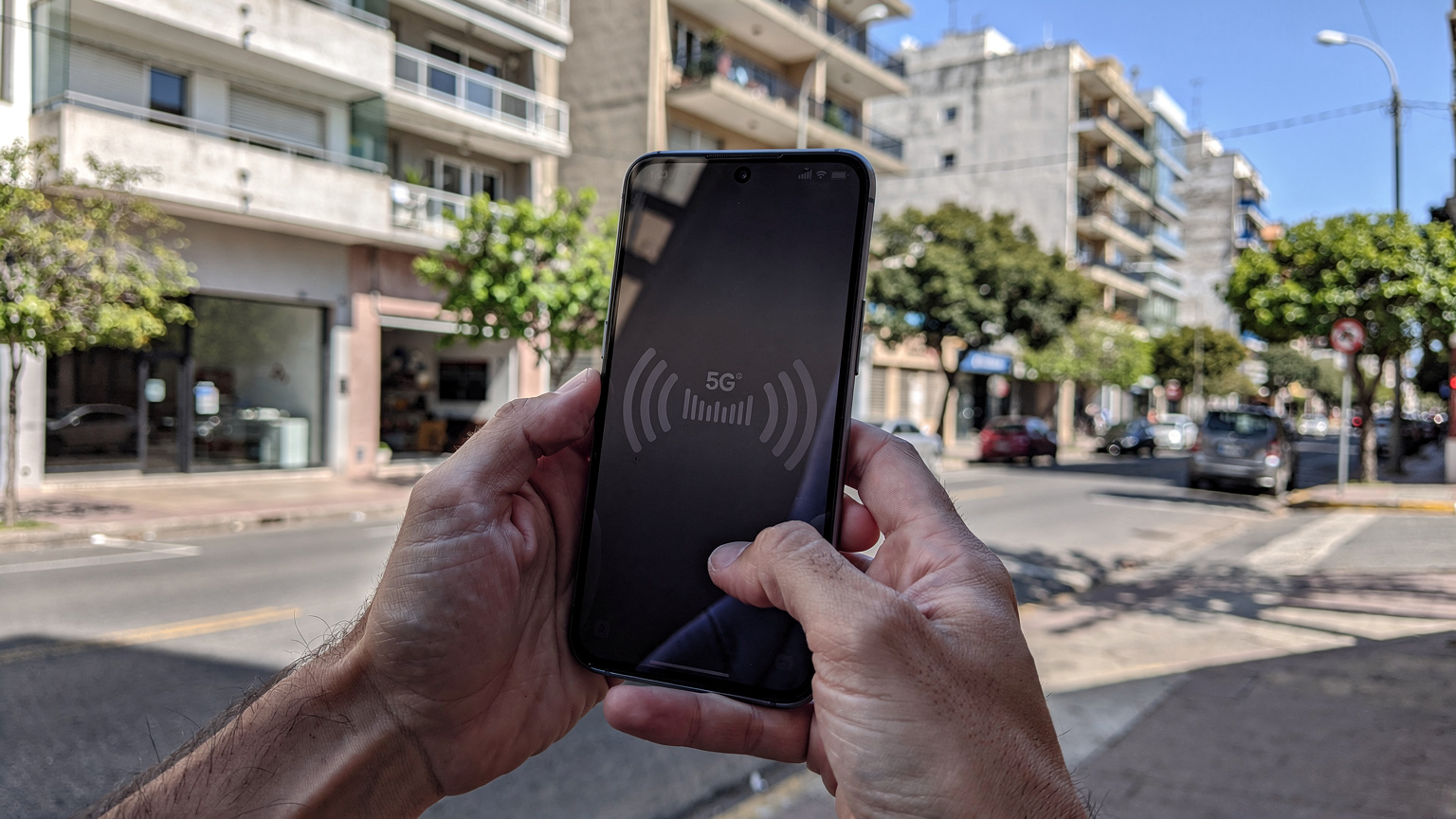 Un smartphone muestra una fuerte señal 5G en una calle concurrida del barrio de Pocitos en Montevideo, con edificios modernos y algunos árboles al fondo, bajo una luz solar intensa de mediodía.