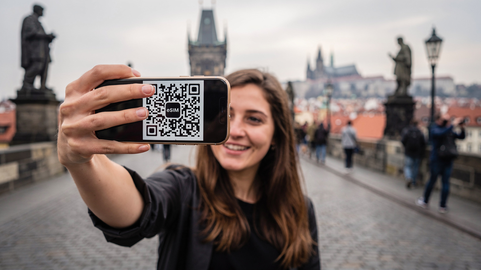 Prag'da tarihi Karl Köprüsü üzerinde, elinde akıllı telefonuyla bir selfie çeken genç bir kadın. Telefon ekranında bir QR kodu belirgin, arka planda şehrin silueti bulanık görünüyor.
