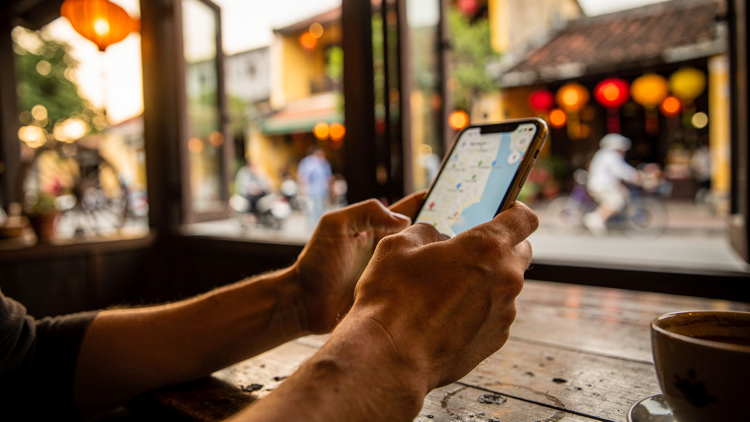 Vietnam'ın Hoi An şehrindeki eski bir kafede, elinde akıllı telefonuyla harita uygulamasını kontrol eden bir gezginin yakın çekim görüntüsü. Arka planda geleneksel fenerler ve hareketli bir sokak silüeti bulanık görünüyor.