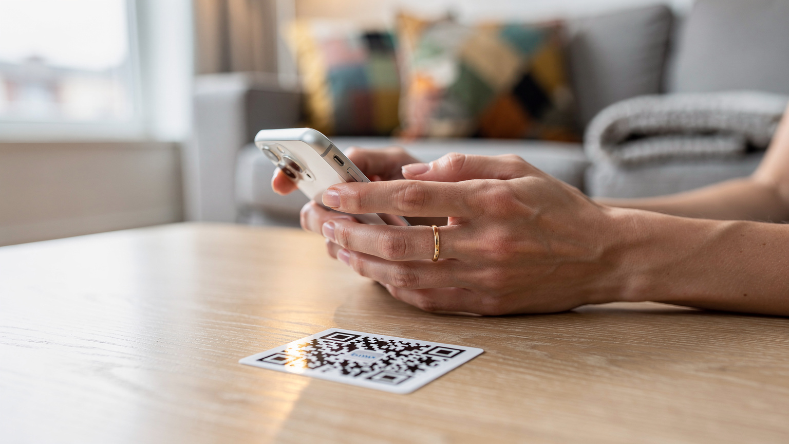 Une main féminine tenant un iPhone en train de scanner un QR code eSIM posé sur une table basse en bois dans un salon lumineux, des coussins colorés et un plaid en arrière-plan. La scène est baignée par la lumière naturelle d'une fenêtre proche.