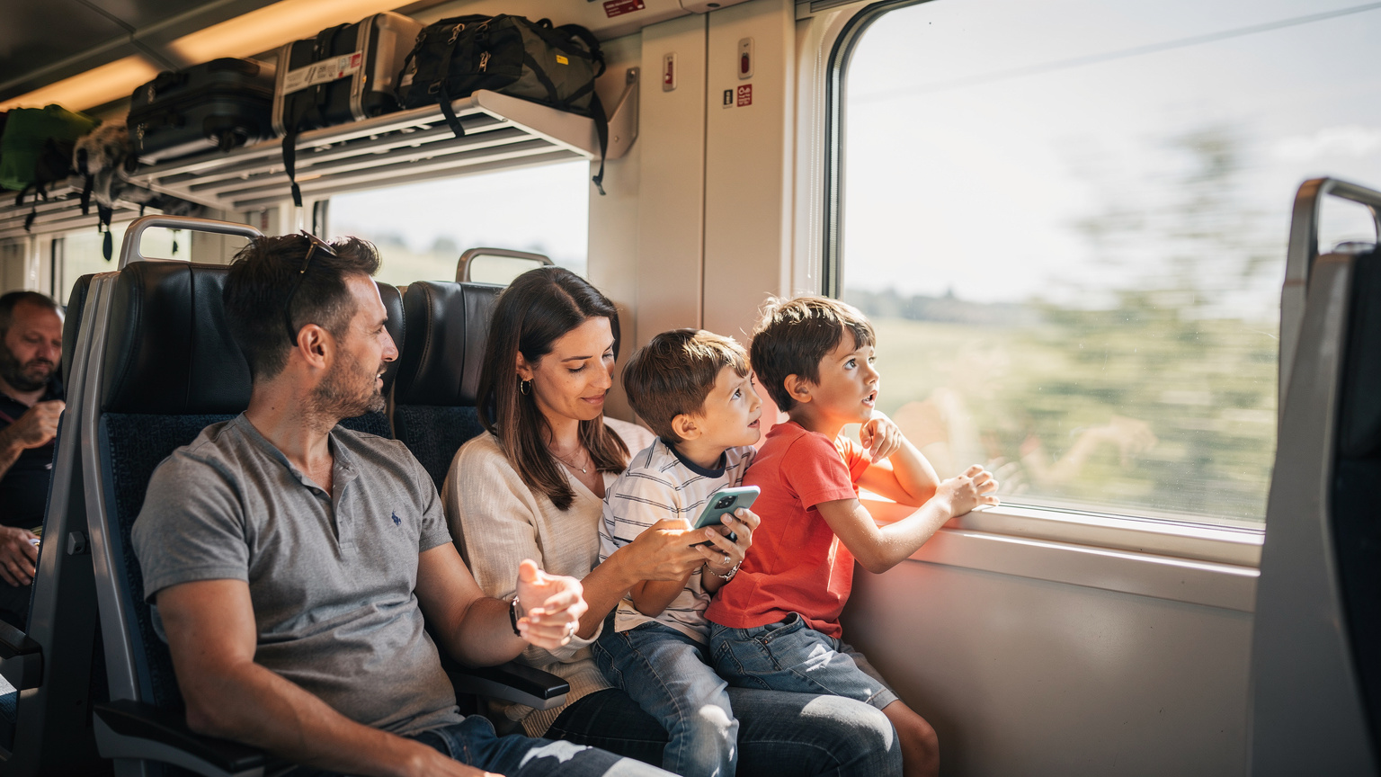 O familie de turiști, formată dintr-un cuplu și doi copii, stau într-un vagon aglomerat al unui tren regional italian. Mama ține un telefon în mână, verificând o hartă offline, în timp ce copiii privesc pe geam. Lumina naturală pătrunde printr-o fereastră mare a trenului.
