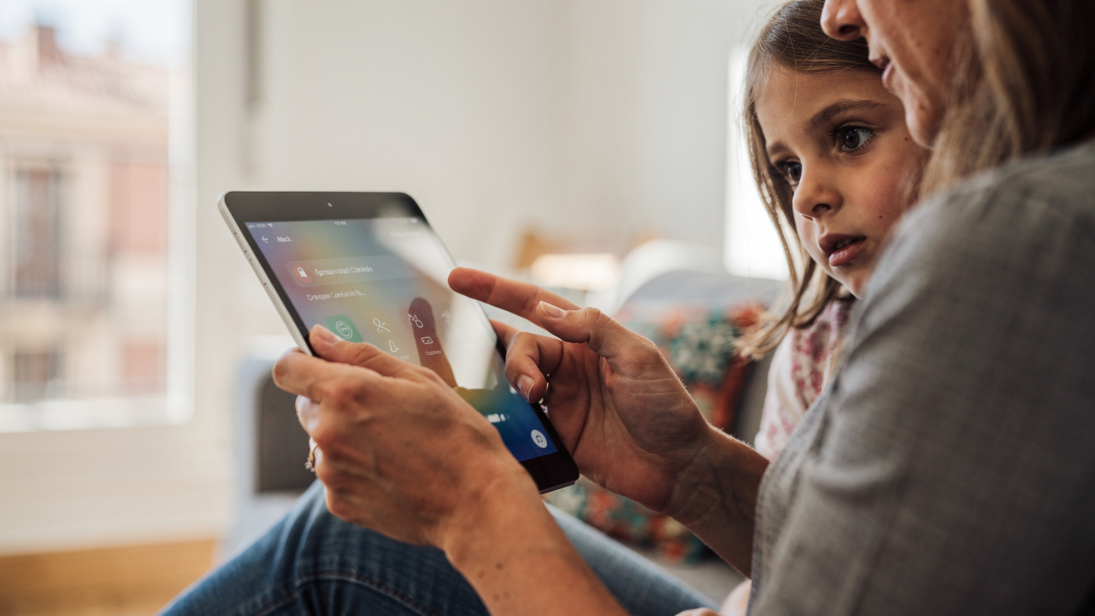 Un primo piano delle mani di una madre italiana che imposta il controllo parentale su un iPad, mentre una bambina di 6 anni curiosa guarda lo schermo da dietro la sua spalla, sedute su un divano in un appartamento vacanze a Barcellona, Spagna, con la luce del mattino che entra da una finestra.