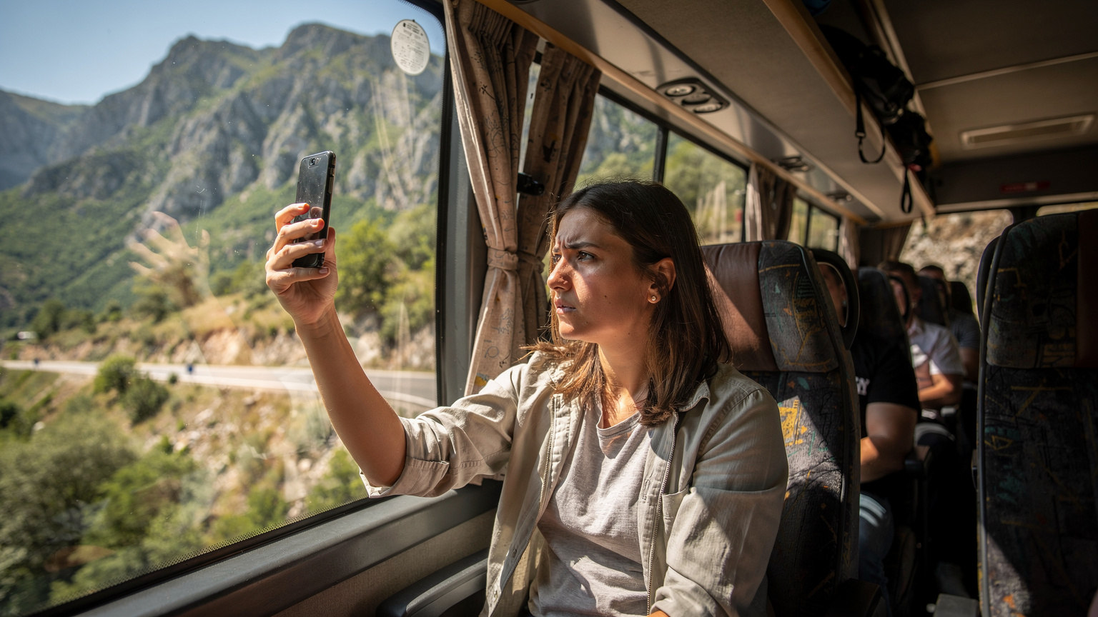 Eine junge Frau sitzt in einem überfüllten Reisebus, der durch eine bergige Landschaft auf dem Balkan fährt. Sie hält ihr Smartphone hoch und versucht, einen schwachen Handyempfang zu finden, während sie besorgt aus dem Fenster schaut.