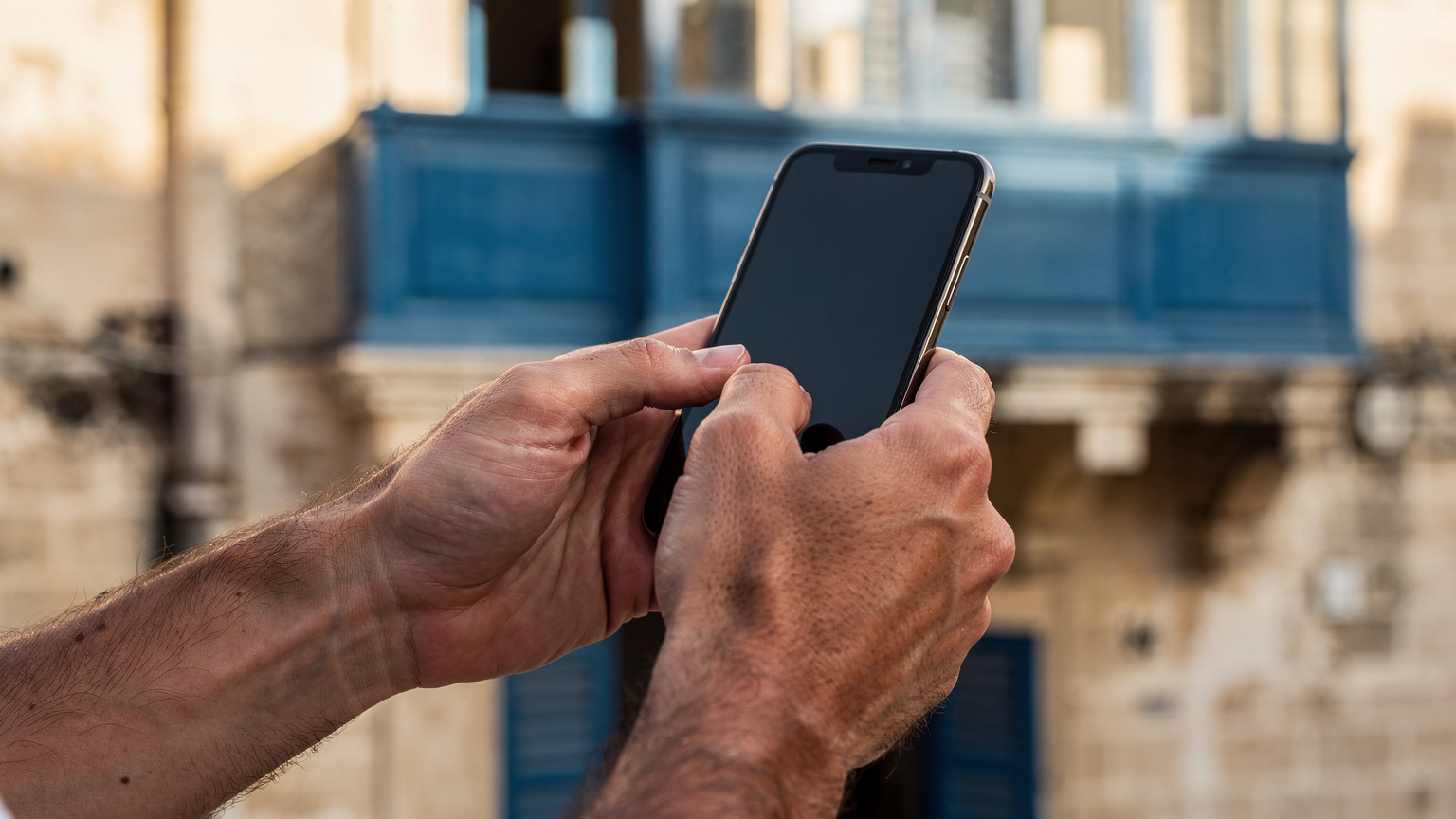 Un primo piano delle mani di una persona che tiene uno smartphone e digita sul display, con una tipica finestra maltese con persiane in legno blu sullo sfondo.