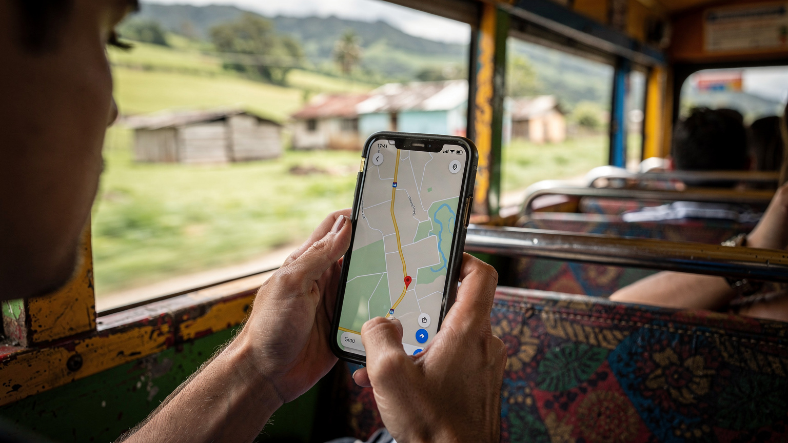 Manos de un viajero sosteniendo un smartphone con una aplicación de mapas abierta, mientras está sentado en un "chicken bus" en movimiento en El Salvador, mirando por la ventana.