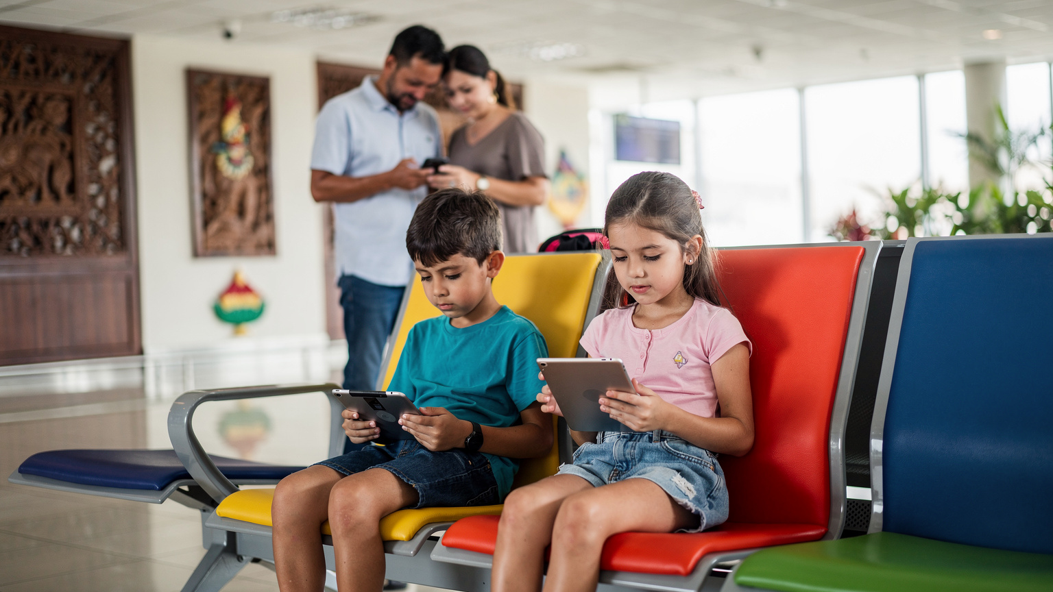 A young boy and girl sitting patiently on a brightly coloured bench at Ngurah Rai International Airport in Bali, each engrossed in a tablet, while their parents check flight details on a smartphone nearby.