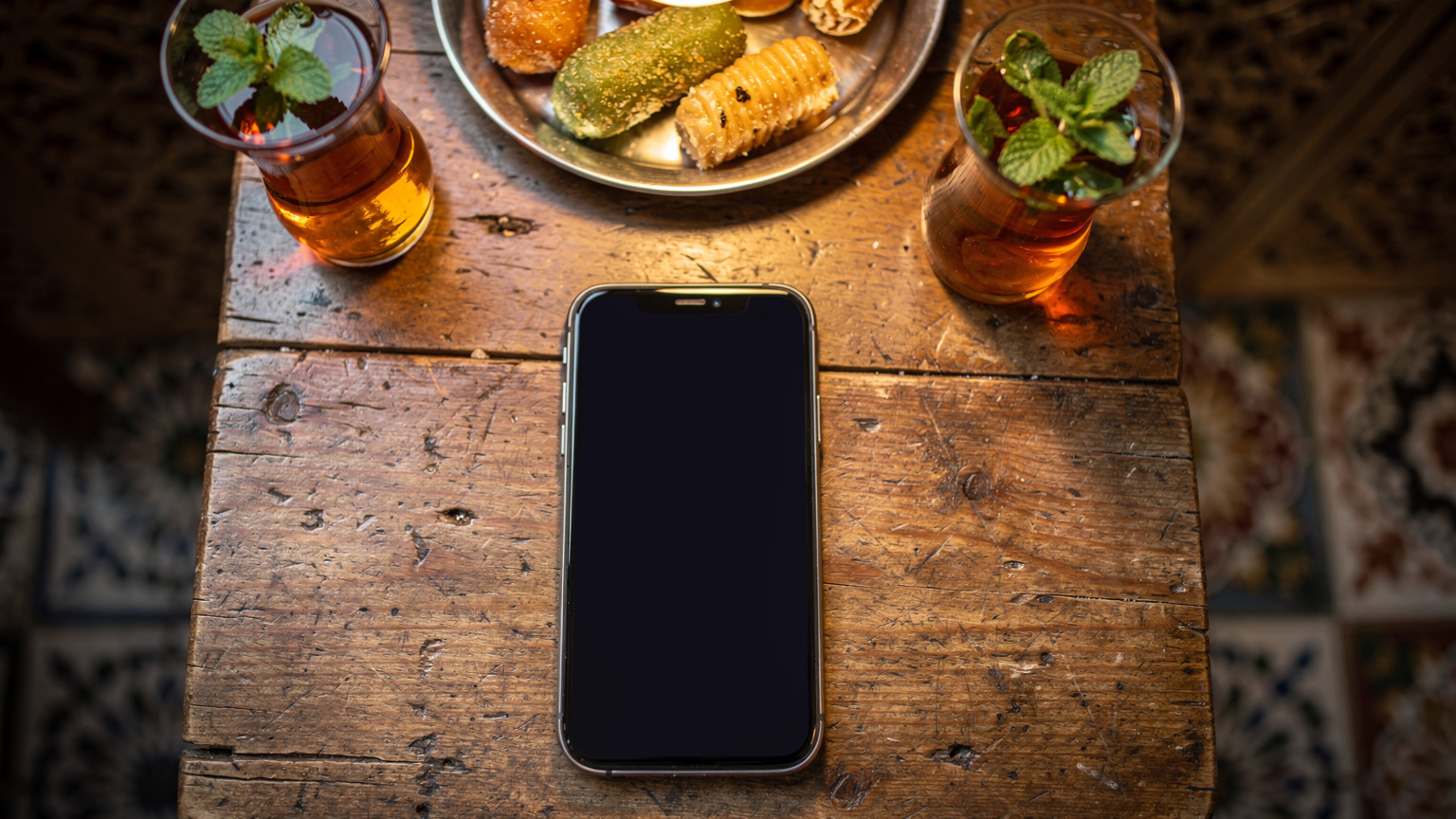 Un smartphone posé sur une table en bois, avec en arrière-plan flou, des tasses de thé à la menthe et des pâtisseries tunisiennes typiques, dans l'ambiance chaleureuse d'un café traditionnel à Tunis.