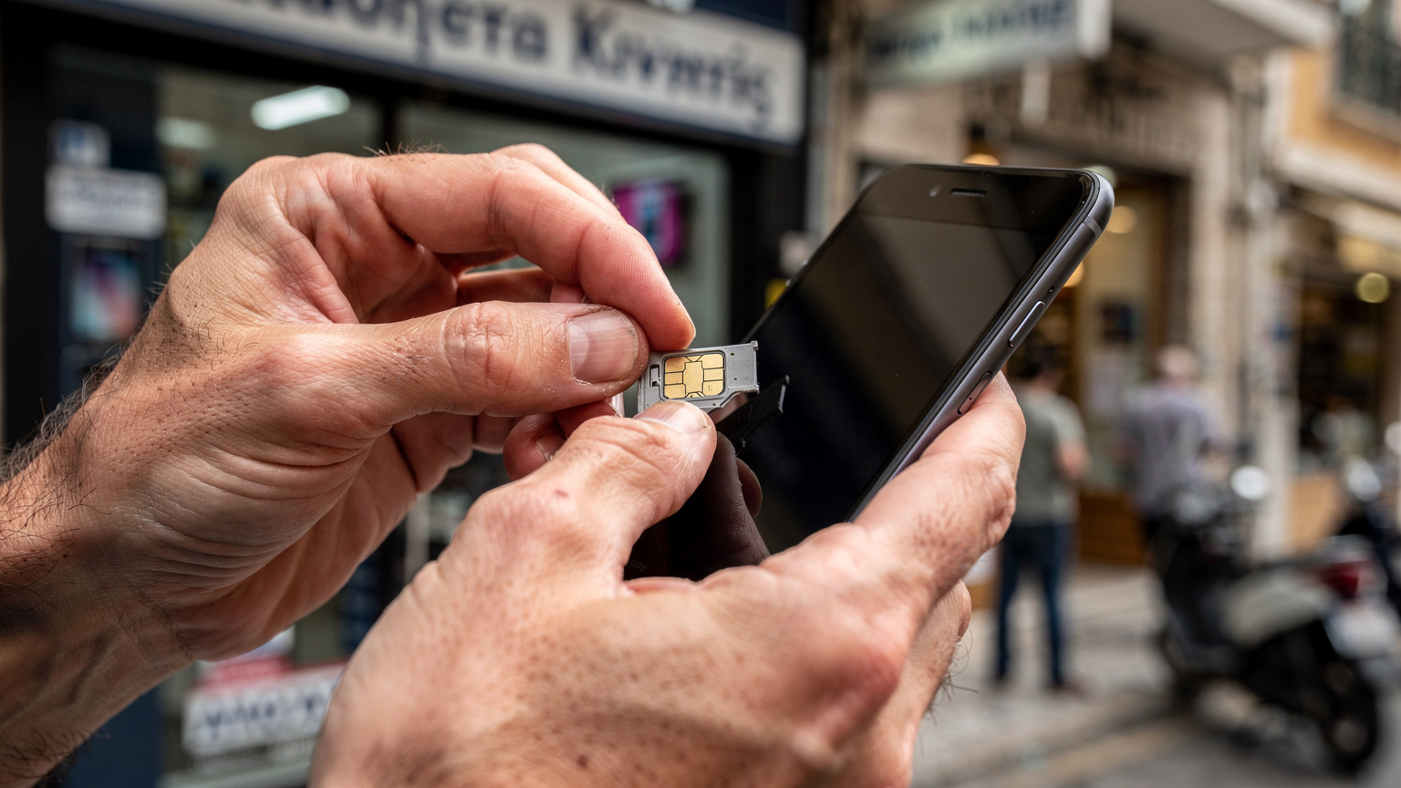 صورة مقربة ليد تضع شريحة SIM فعلية في درج هاتف ذكي، مع خلفية غير واضحة لمحل اتصالات في شارع مزدحم بأثينا.