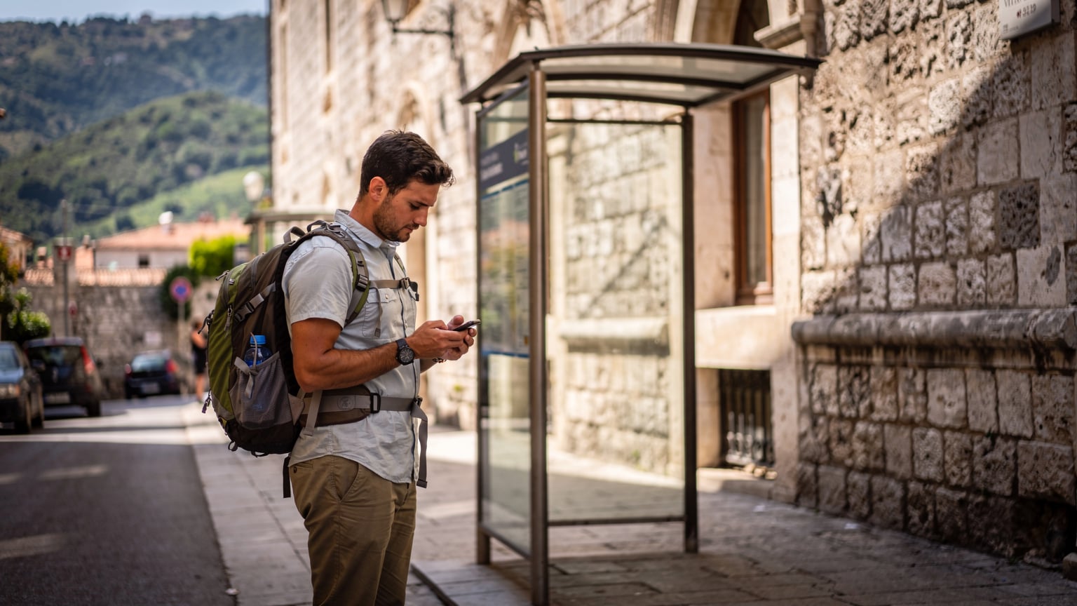 Un turista con uno zaino sta consultando il suo telefono mentre aspetta alla fermata dell'autobus, con una facciata di edificio storico sullo sfondo, in una giornata soleggiata.