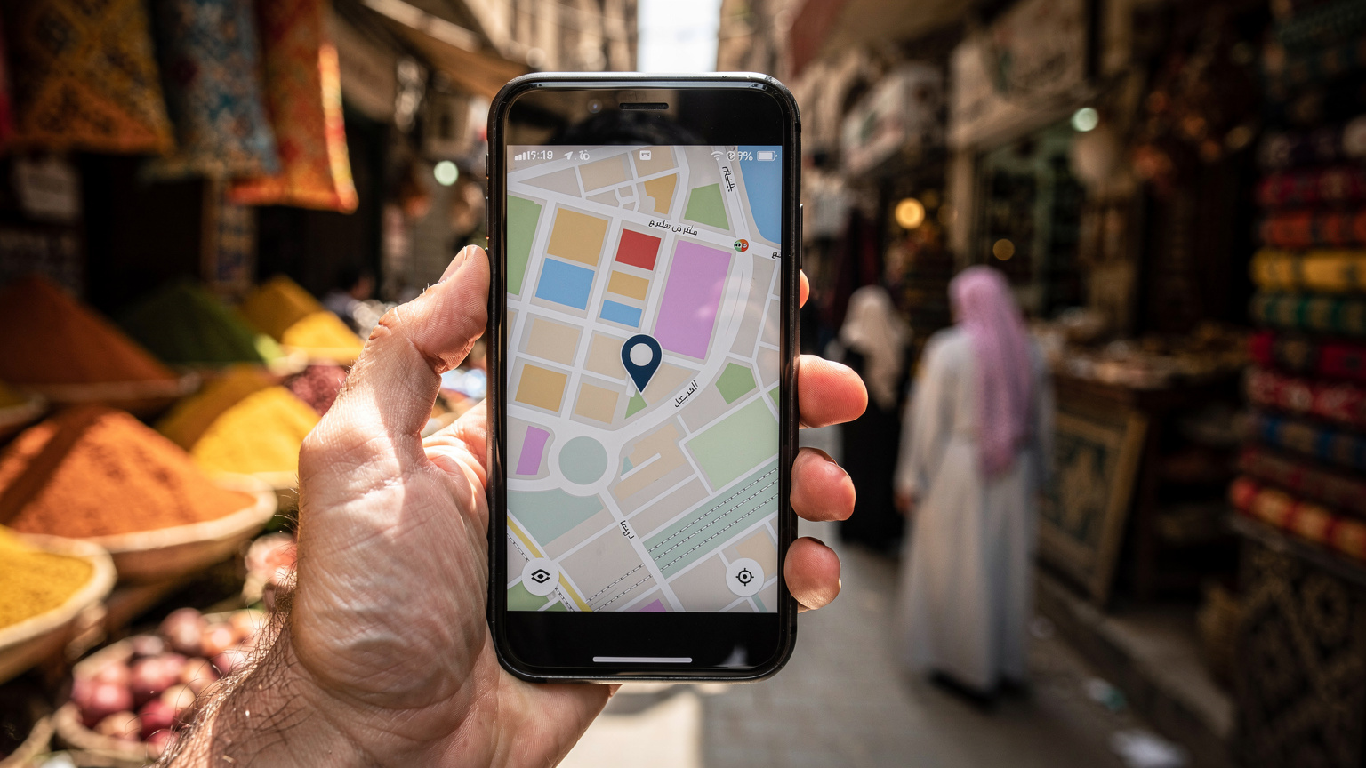 صورة مقربة ليد تمسك بهاتف ذكي يعرض تطبيق خرائط، مع خلفية غير واضحة لسوق شعبي صاخب في مدينة القاهرة القديمة.