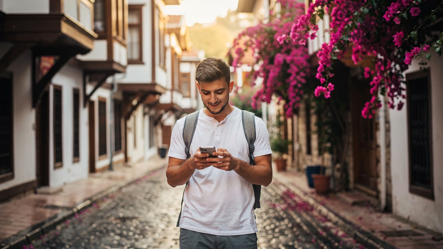A tourist checks his phone amidst the narrow, historic streets of Kaleiçi in Antalya, with traditional Ottoman-style houses lining the cobblestone path.