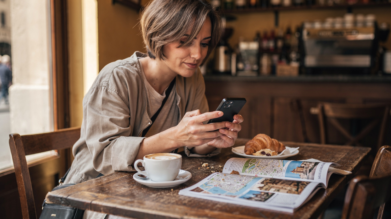 O călătoare își activează eSIM-ul pe telefon la o masă de cafenea din Florența, cu o cană de cappuccino și un ghid turistic deschis pe masă.