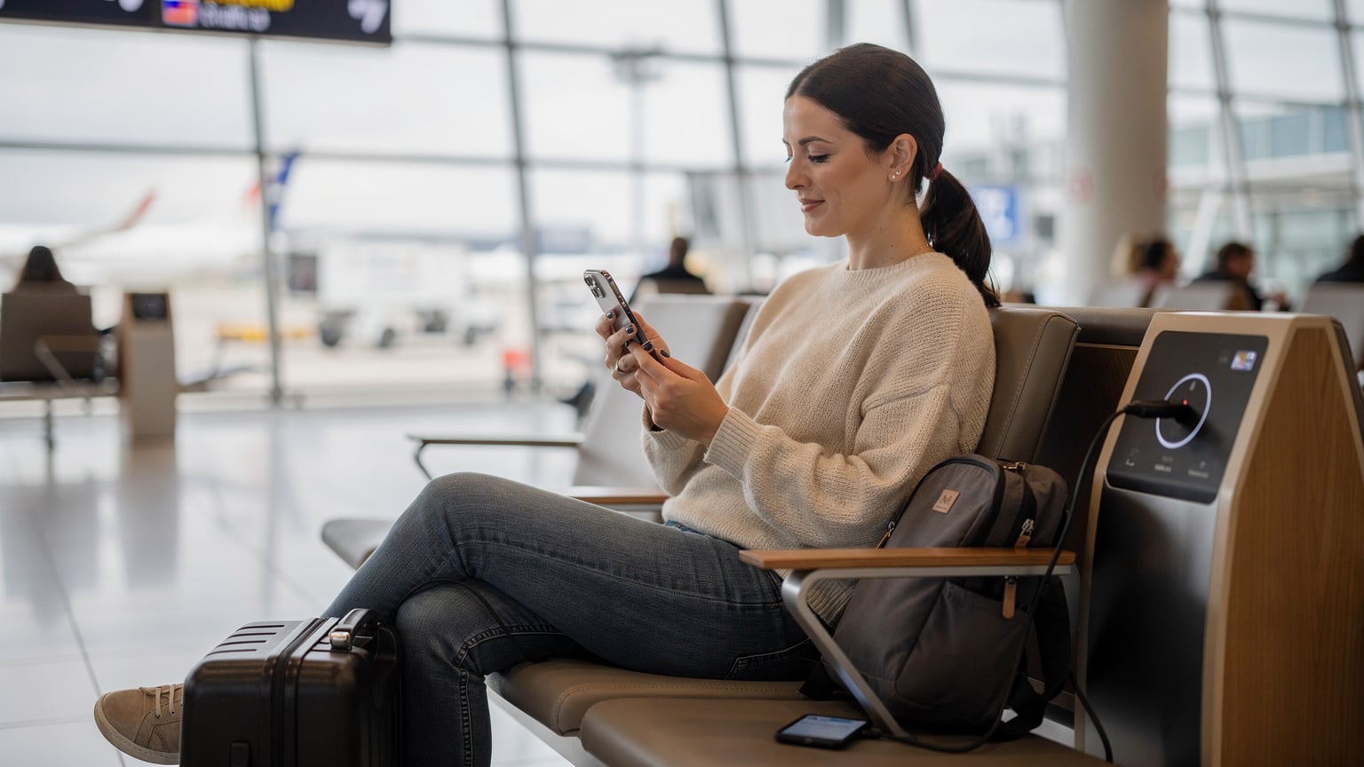 A woman at a departure gate in a French airport, looking at her smartphone with a travel app open, verifying her Cellesim eSIM details before boarding her flight to Indonesia.