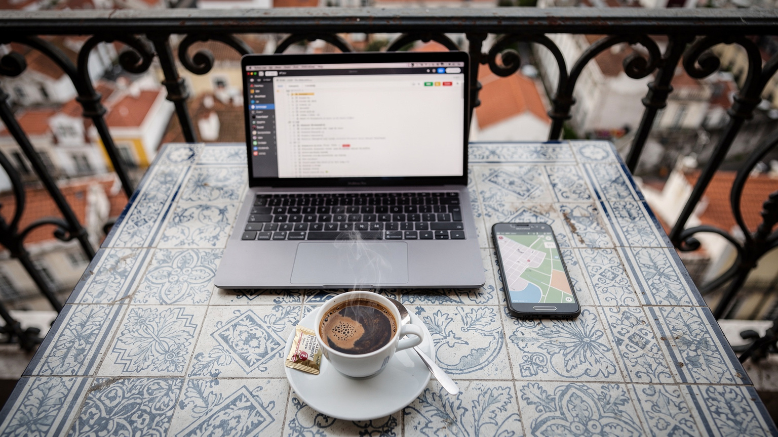 Uma mesa de café em Lisboa, Portugal, com um laptop, um smartphone exibindo um mapa, e uma xícara de café.