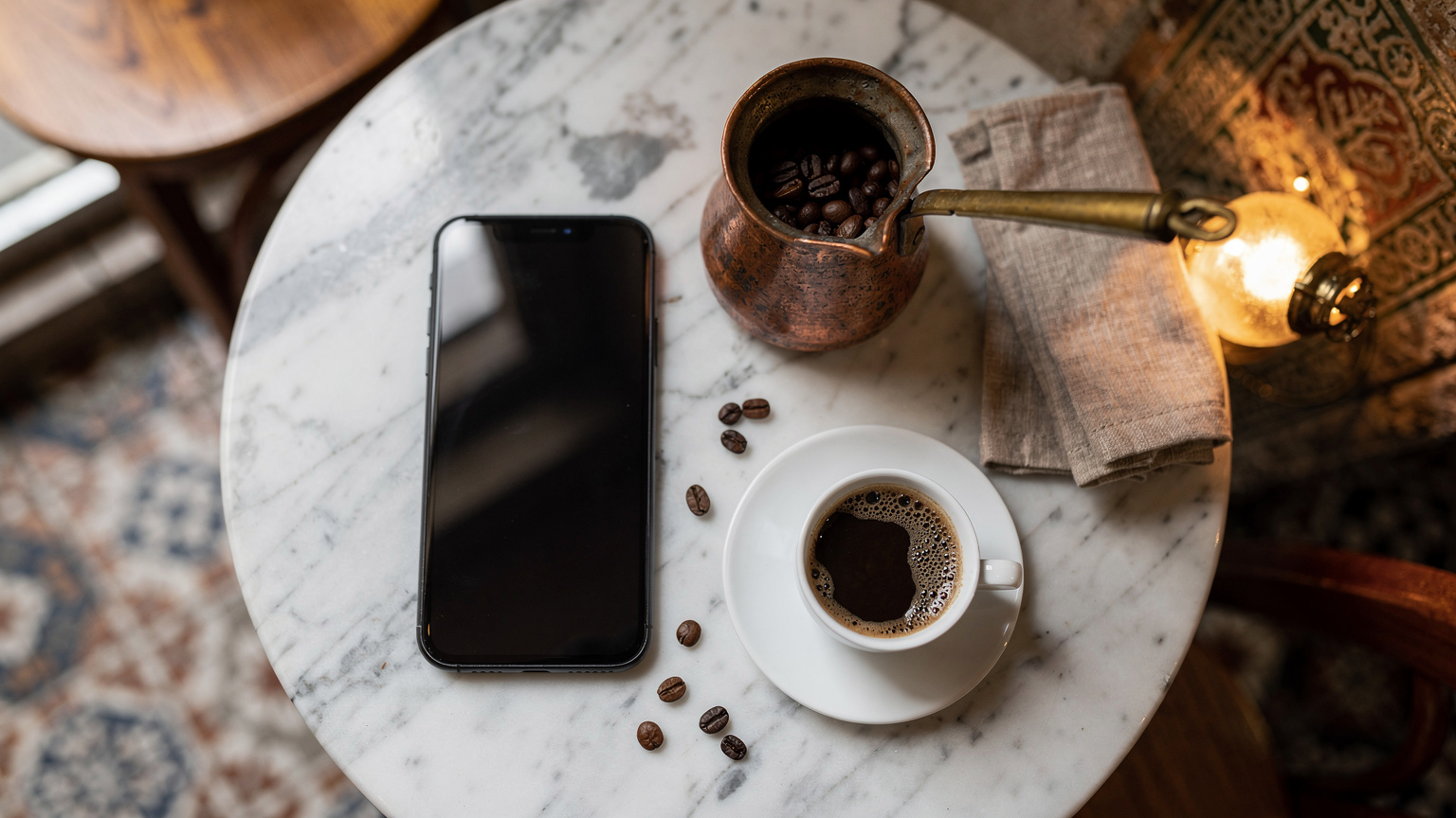 Geleneksel bir Türk kahvehanesinde, mermer bir masanın üzerinde, el yapımı bakır bir cezve ve fincanın yanında bir akıllı telefon zarifçe duruyor.