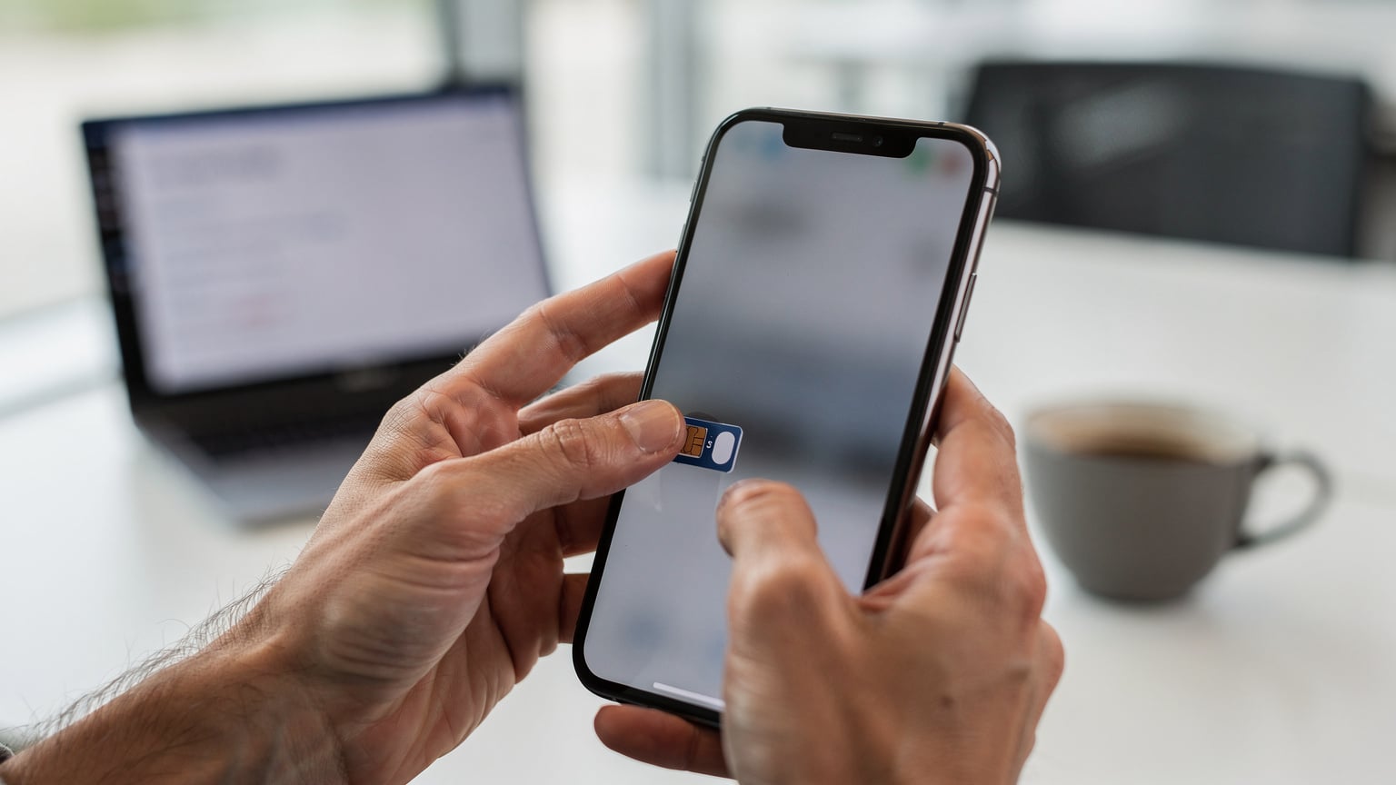 A close-up of a person's hands holding an iPhone in a modern co-working space, demonstrating how to switch between cellular plans in the settings, with a laptop screen subtly blurred in the background.