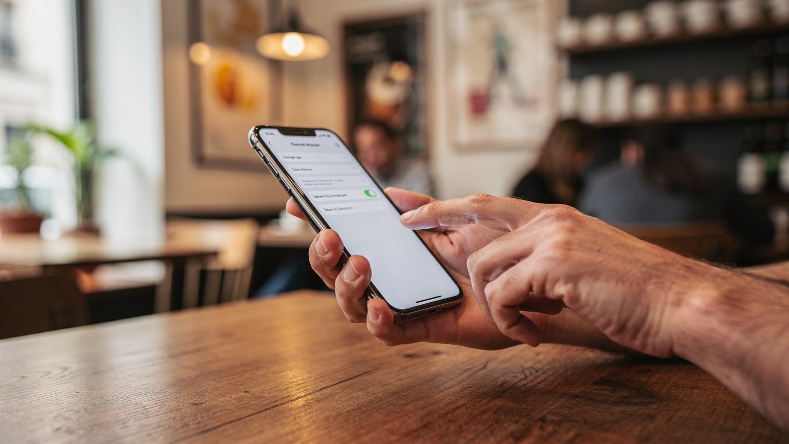 A traveler in a comfortable European cafe, looking at their iPhone and carefully navigating the settings menu to configure their eSIM for data and ensure their physical SIM remains active for SMS.