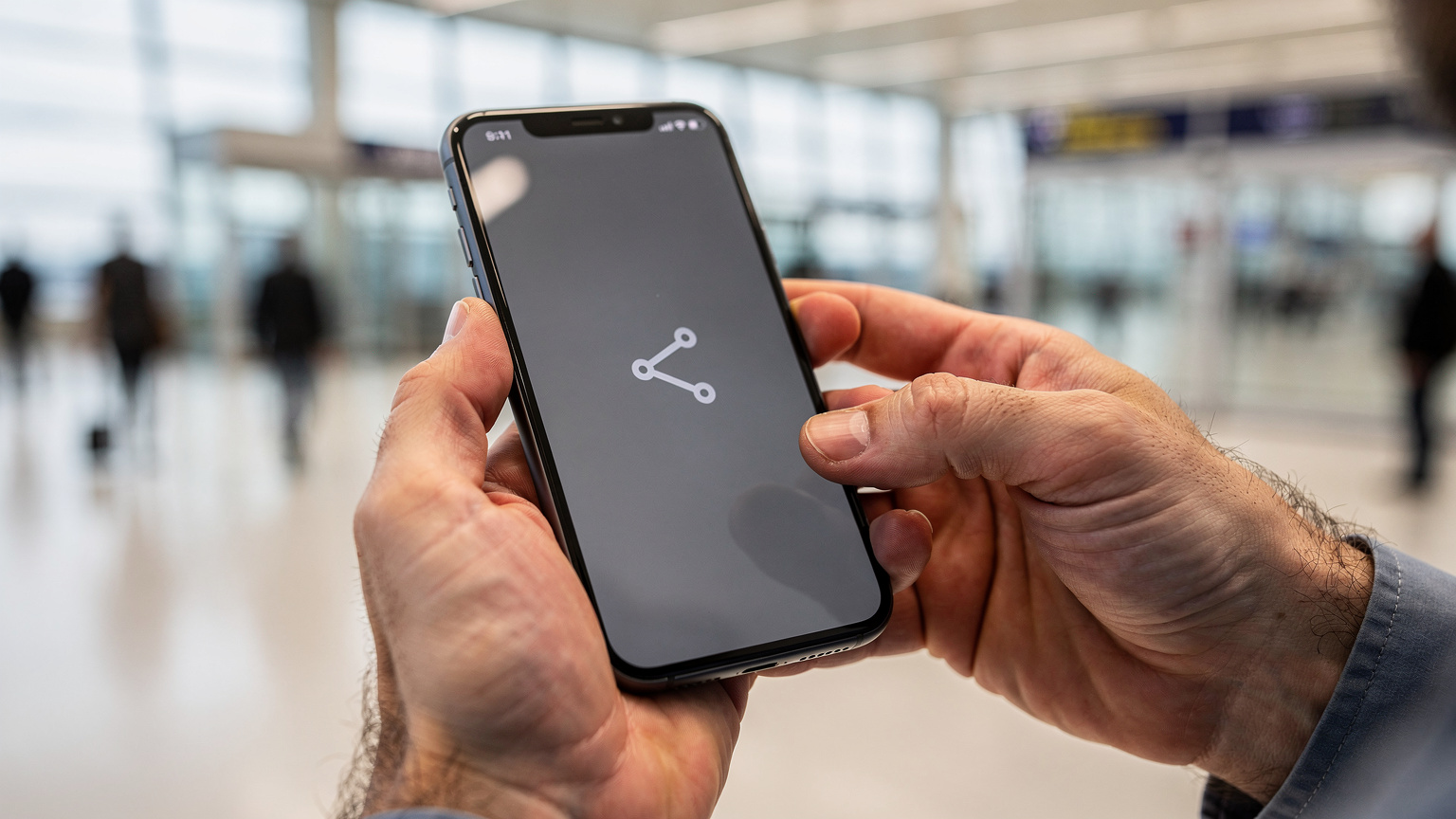 Detailní záběr na mužské ruce, které drží smartphone a prstem klepají na obrazovku, kde je vidět ikona nastavení sítě. Scéna se odehrává v interiéru moderního letištního terminálu, v pozadí je rozmazaná letištní hala plná cestujících.