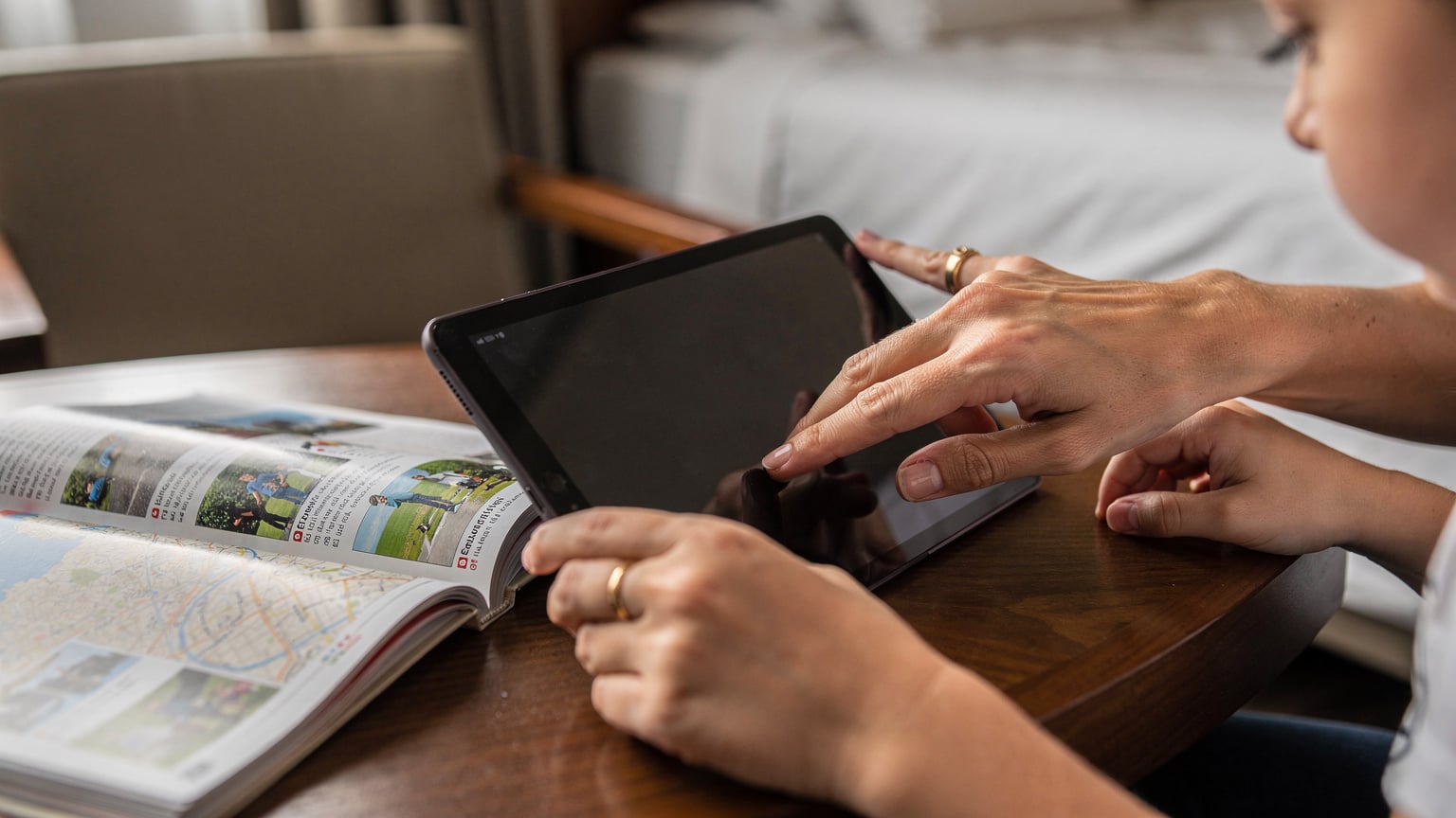 A close-up shot of a woman's hands, gently helping a child navigate a tablet. The child's device shows a map application, indicating active use for navigation.