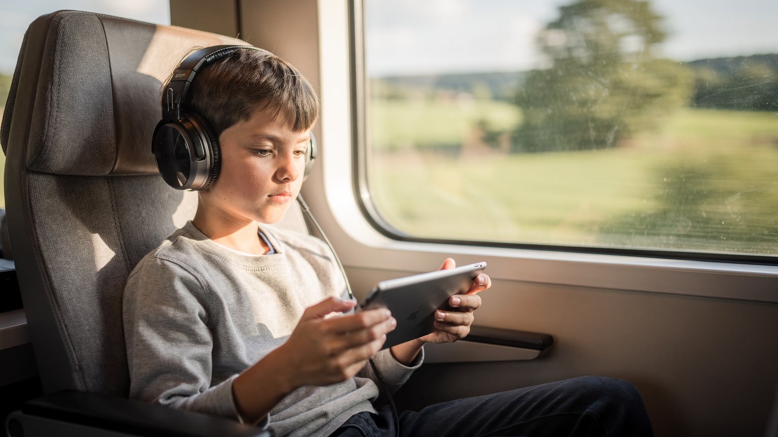 A child, around 9 years old, engrossed in a tablet game while sitting comfortably in a brightly lit train carriage, passing through a picturesque European countryside.