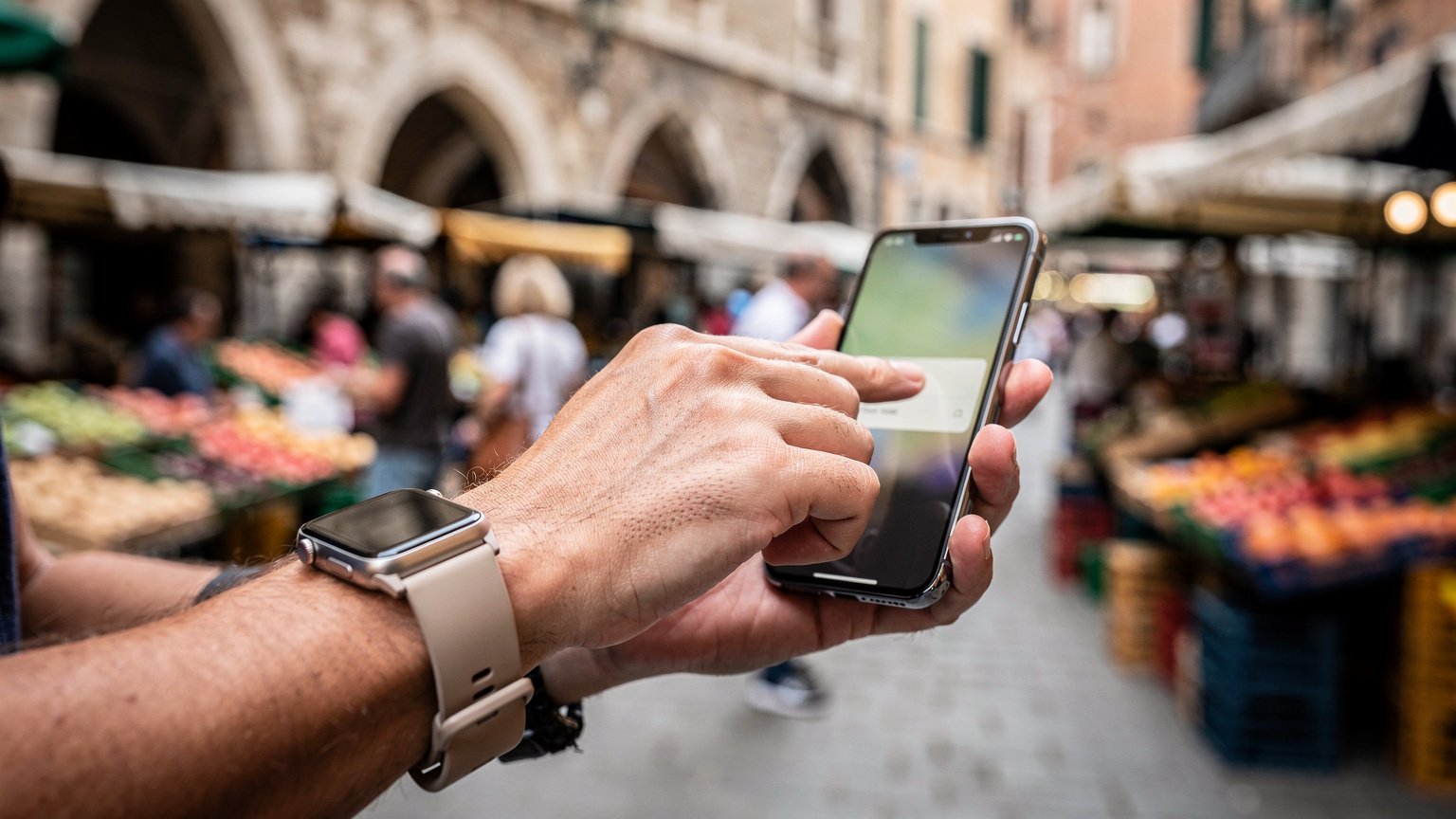 O mână de părinte, purtând un ceas inteligent elegant, ține un smartphone, ajustând setările de control parental pe ecranul dispozitivului, în timp ce în fundal se zărește o piață plină de viață dintr-un oraș european, cu tarabe și oameni trecând.