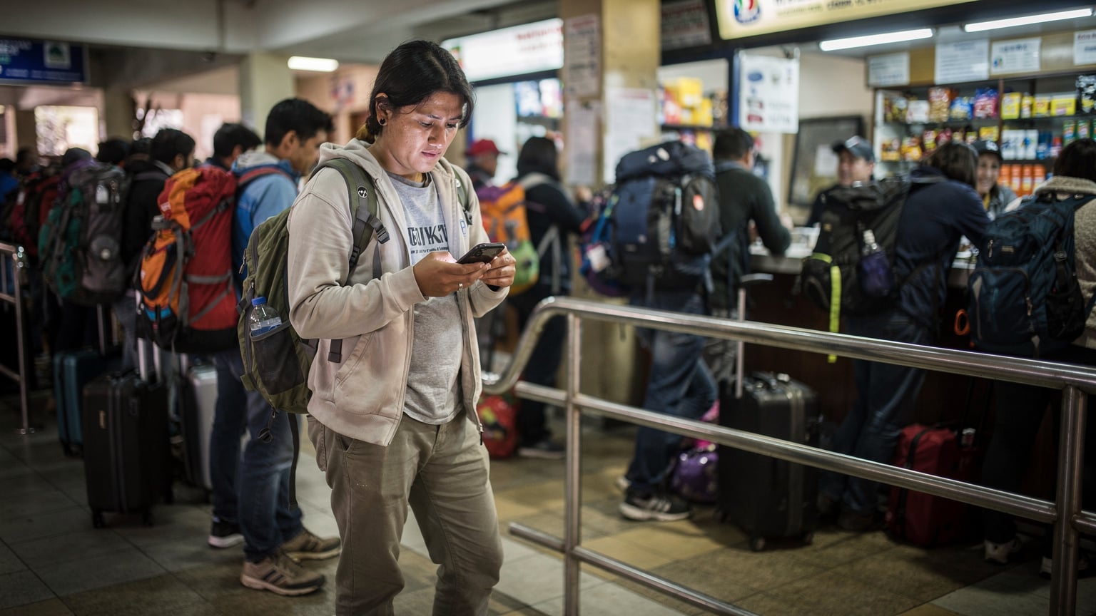 A person standing in line at a busy bus terminal in La Paz, Bolivia, looking at their phone with a slightly concerned expression, trying to confirm a bus schedule or ticket details, surrounded by other travelers and luggage.