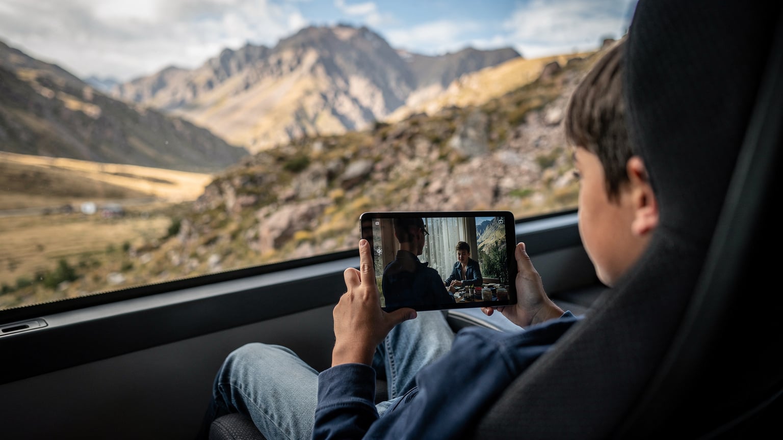 A child's hands holding an iPad displaying a downloaded movie, seen from over their shoulder on a long-distance bus journey through the Chilean countryside.