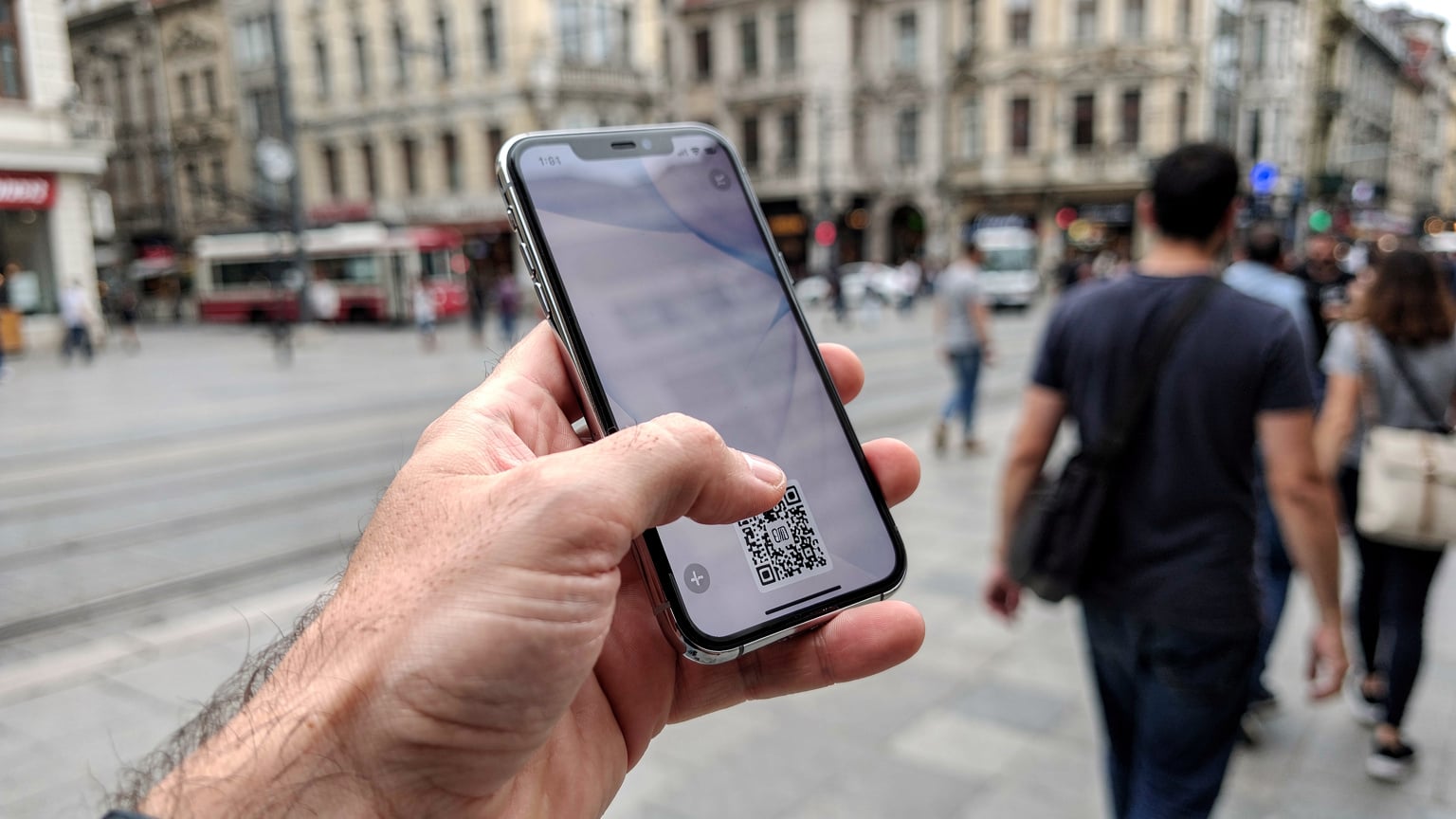 Yurt dışı seyahatlerinde, telefon ekranında bir eSIM QR kodu tarama işlemi sırasında bir elin akıllı telefon tuttuğu yakın çekim fotoğrafı. Arka planda hareketli bir şehir meydanı hafifçe bulanık görünüyor.