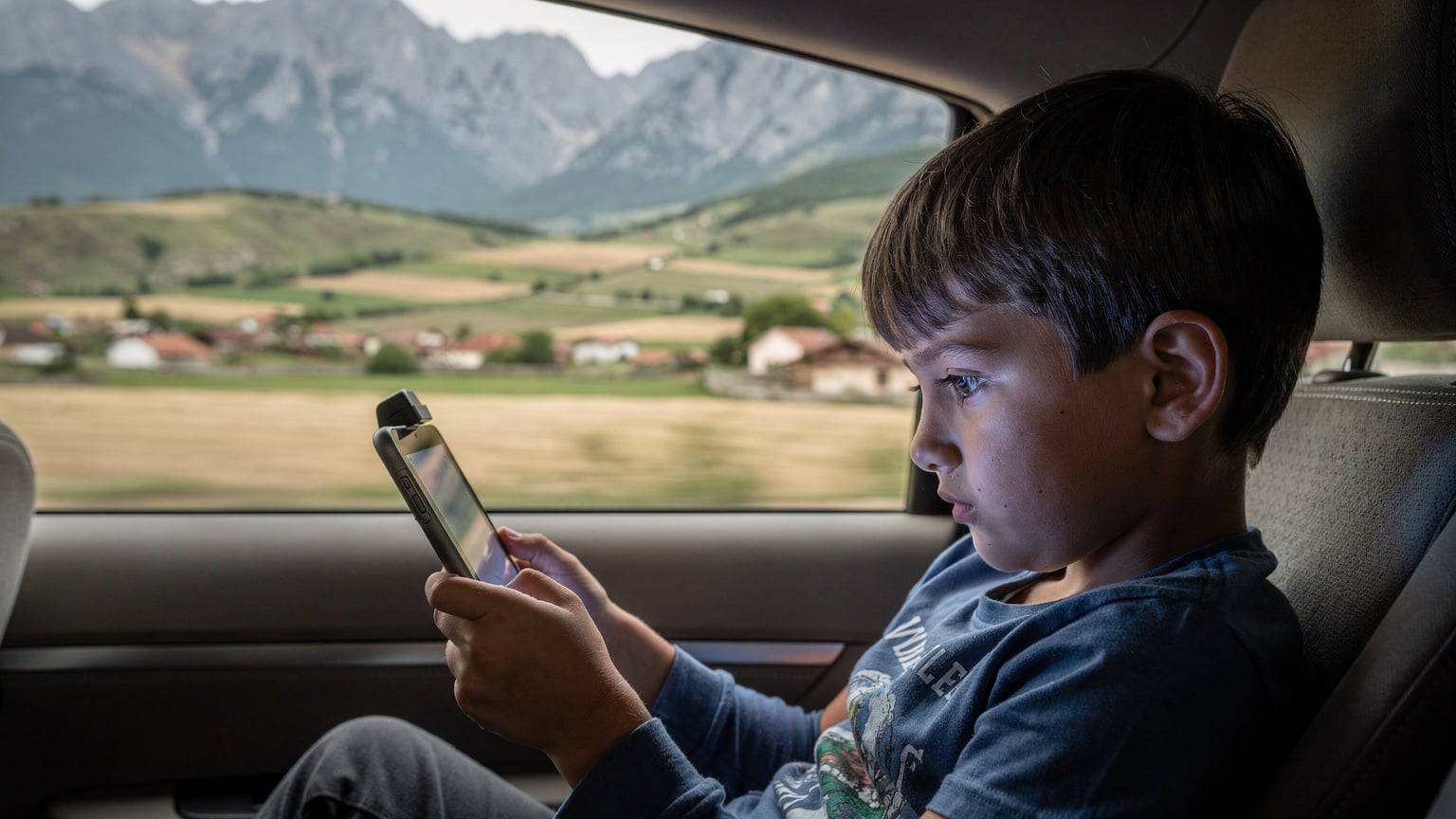 Ein Kind im Alter von 10 Jahren sitzt auf der Rückbank eines Autos und schaut auf ein Tablet, das an einer Kopfstütze befestigt ist, während die Landschaft Nordmazedoniens durch das Fenster zieht.