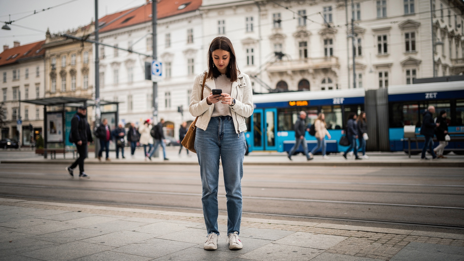 Een drukke bushalte in het centrum van Zagreb, Kroatië, met mensen die wachten op de ZET-tram, terwijl een jonge vrouw geconcentreerd naar haar telefoon kijkt om een route te controleren.