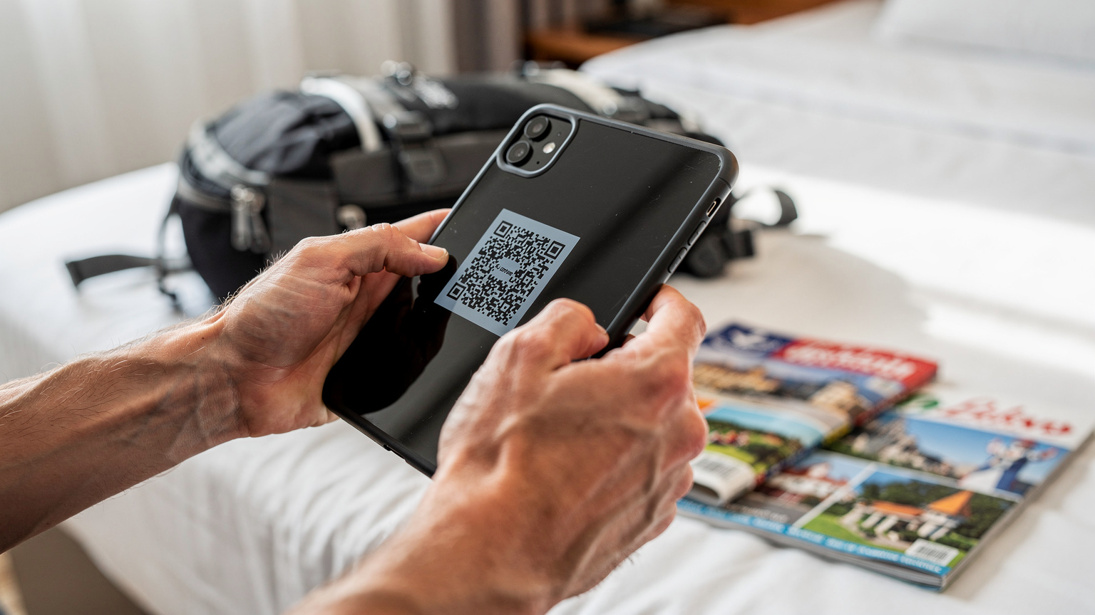 Close-up van handen die een smartphone vasthouden en een QR-code scannen voor eSIM-activatie in een lichte hotelkamer in Belgrado, Servië, met een rugzak en een reismagazine op de achtergrond.