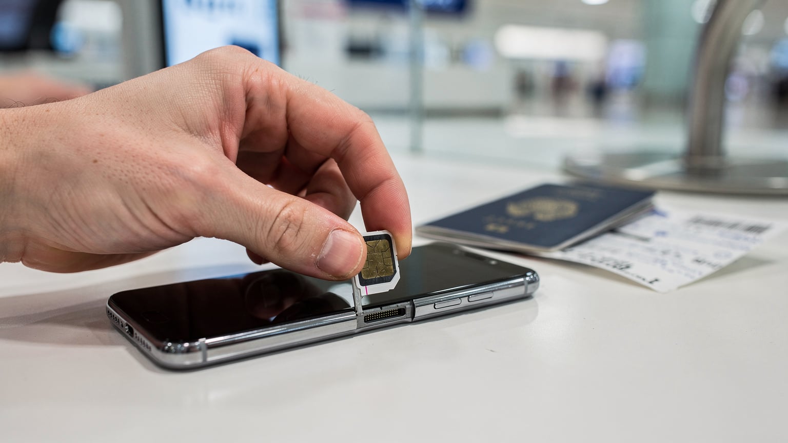 A close-up of a traveller's hands carefully inserting a physical SIM card into a smartphone at a brightly lit counter in a phone store at Incheon Airport, Seoul, with travel documents slightly blurred in the background.