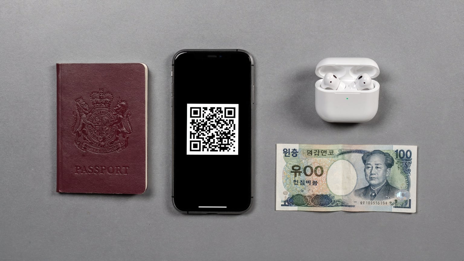 An editorial overhead flat-lay shot of a passport, a smartphone displaying an eSIM activation QR code, a pair of wireless earbuds, and a Korean Won banknote, all arranged on a modern grey desk surface, suggesting preparation for international travel.