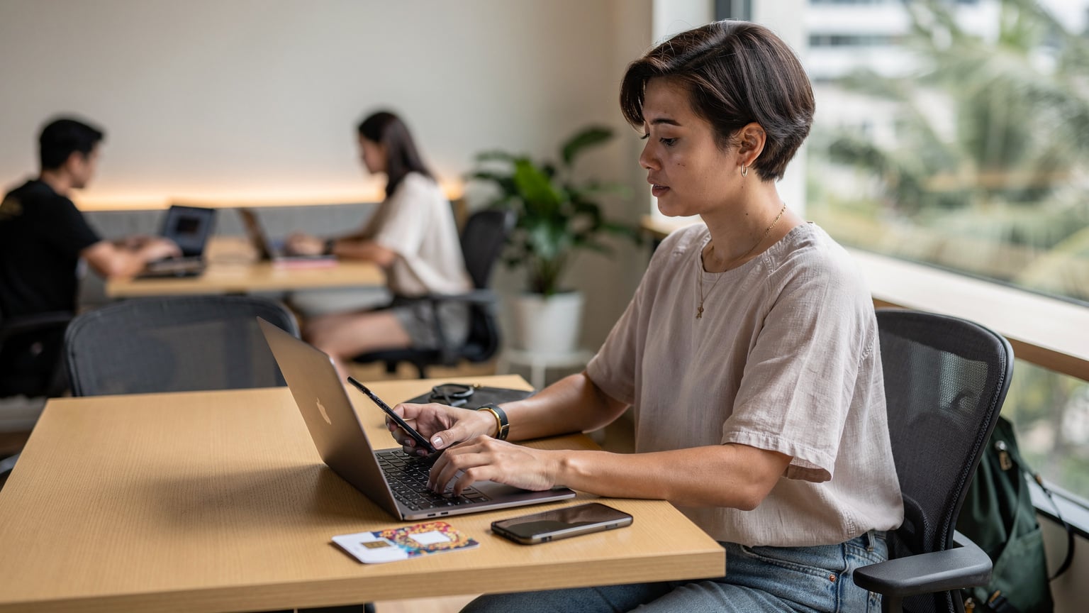 タイ・バンコクのThe Hiveコワーキングスペースで、ノートPCを開きながらスマートフォンを操作しているフリーランサーの女性。手元にはタイのSIMカードが置かれている。