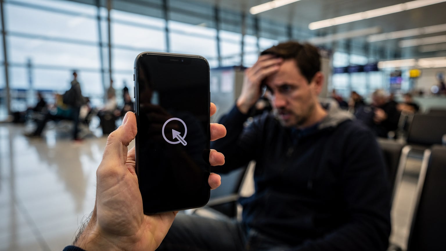 Eine Nahaufnahme eines Smartphones, das in der Hand gehalten wird, mit der Anzeige einer Fehlermeldung auf dem Bildschirm, während sich eine Person besorgt die Stirn reibt. Die Umgebung ist ein überfüllter, heller Wartebereich eines Flughafens.