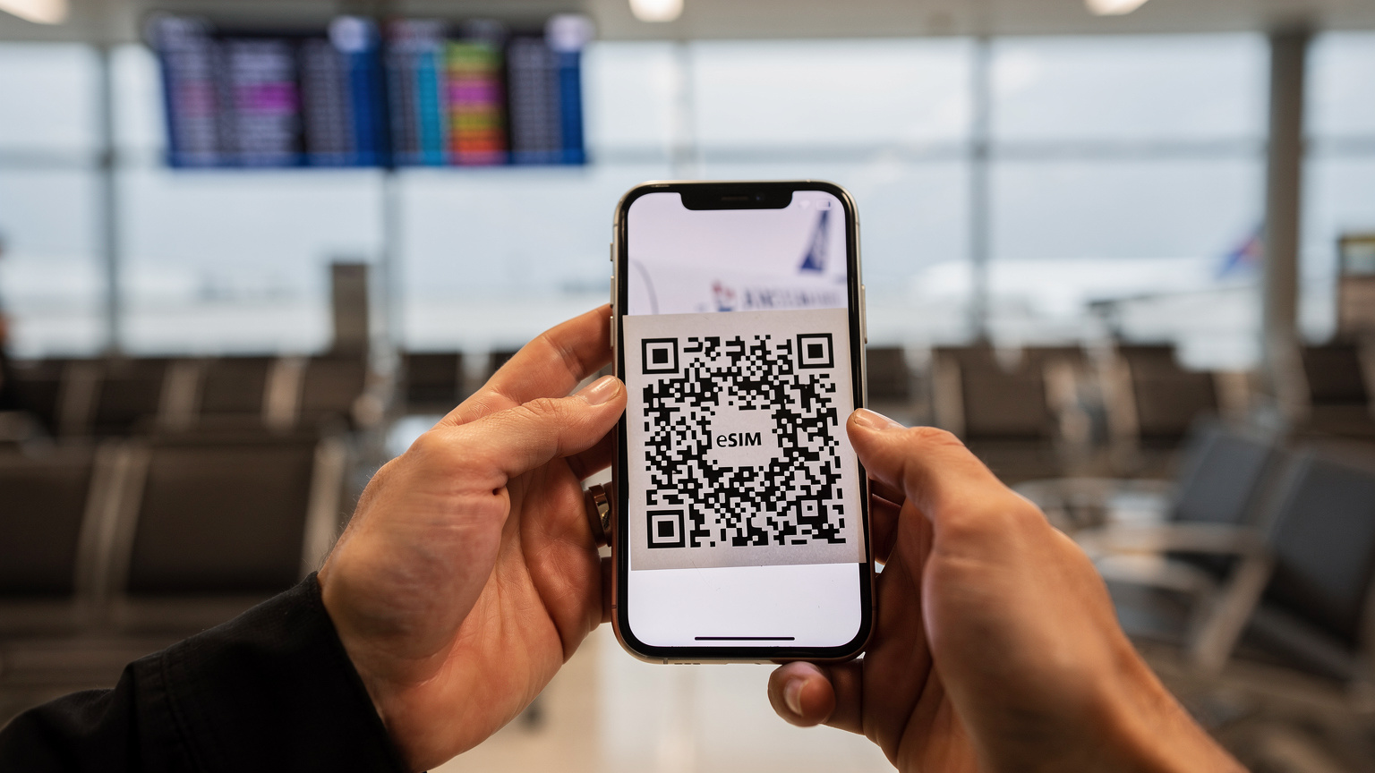 Närbild på en resenärs händer som håller en smartphone och skannar en QR-kod för eSIM-aktivering, med en flygplatsgate i oskärpa i bakgrunden.