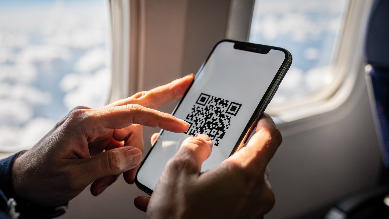 A close-up of a traveler's hands holding an iPhone, scanning a QR code for eSIM activation, with a blurred airplane window and cloudscape in the background, suggesting activation mid-flight.