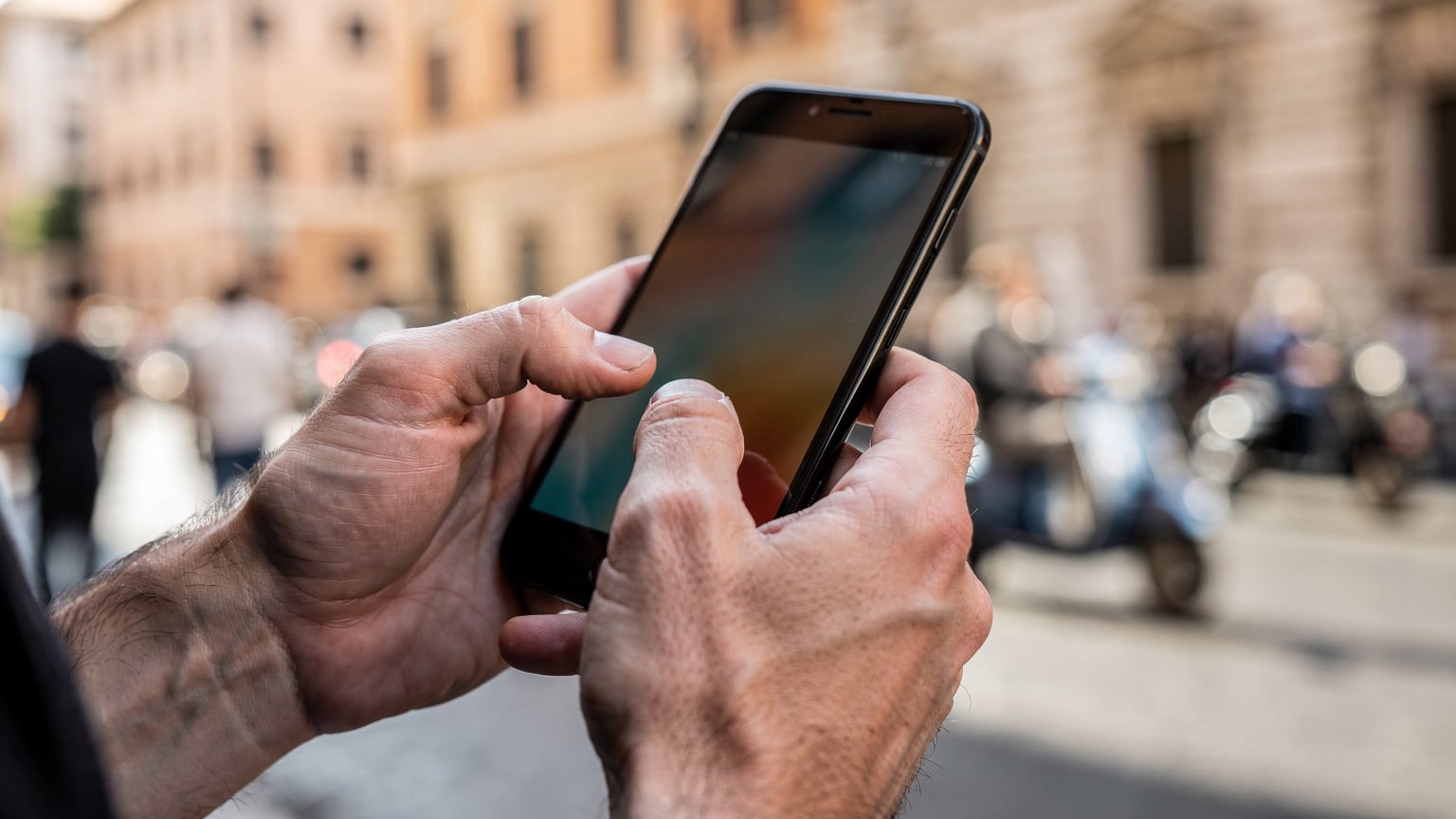 Un primo piano delle mani di una persona che tiene uno smartphone, mostrando l'interfaccia di attivazione di un'eSIM, con uno sfondo leggermente sfocato di una strada trafficata di Roma.
