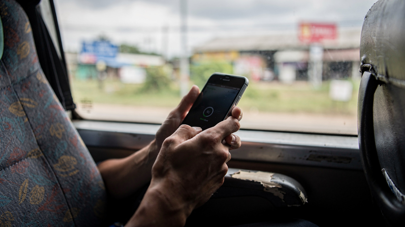 Tangan seorang pengembara memegang telefon pintar dengan skrin menunjukkan notifikasi 'No Service' atau 'Roaming', di dalam bas yang baru saja melintasi sempadan darat, dengan pemandangan luar tingkap yang berubah.