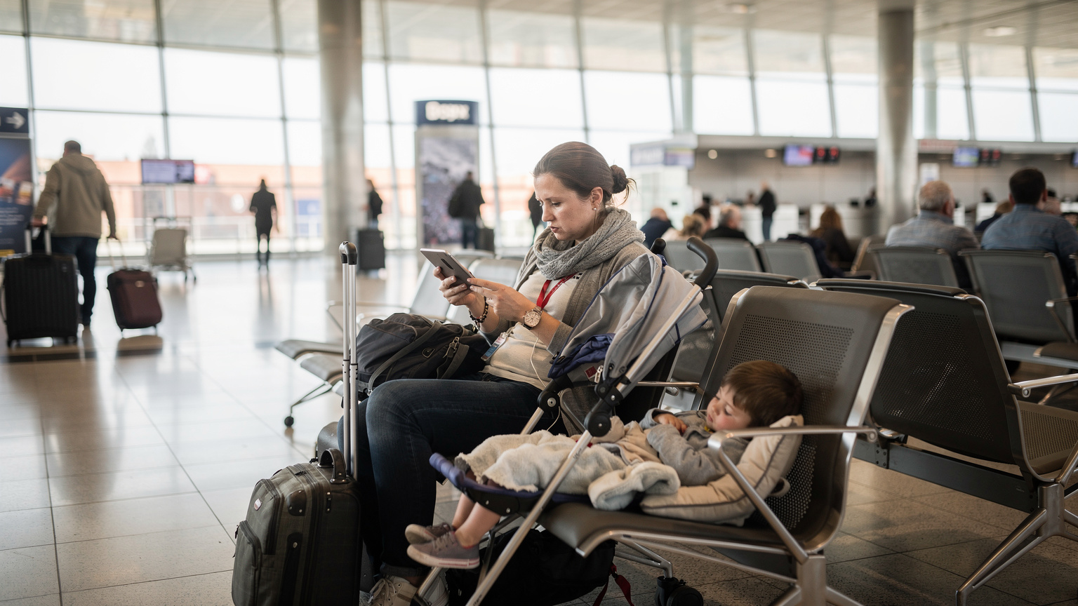 Cestovatelka se snaží připojit k Wi-Fi v přeplněné letištní hale v Praze, s únavou v očích a tabletem v ruce, zatímco její dítě spí v kočárku.