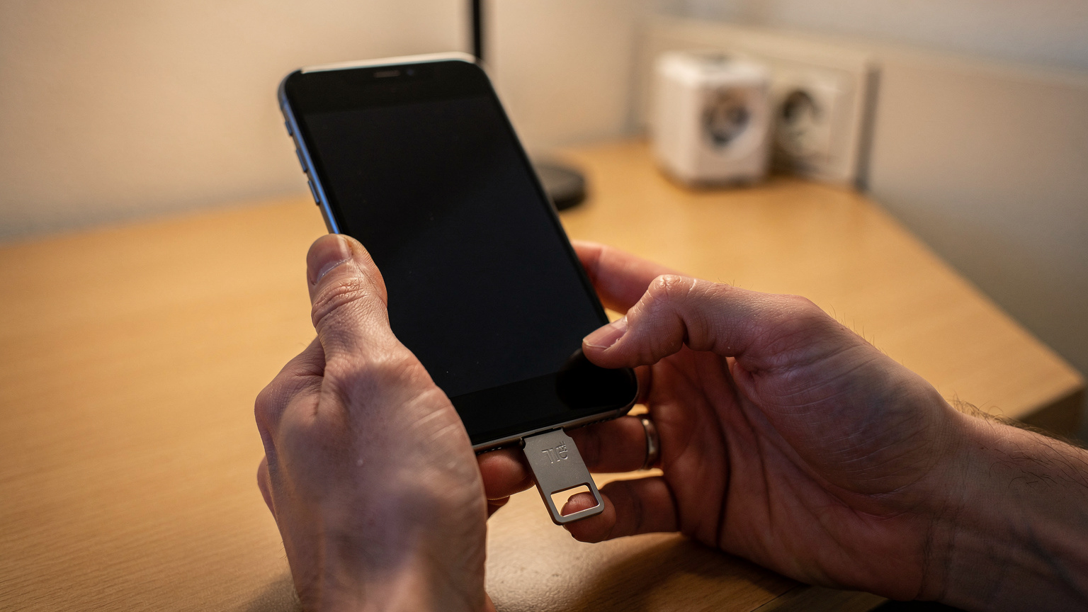 A person's hands holding an iPhone with a hardware security key plugged into it, on a hotel room desk with a travel adaptor.
