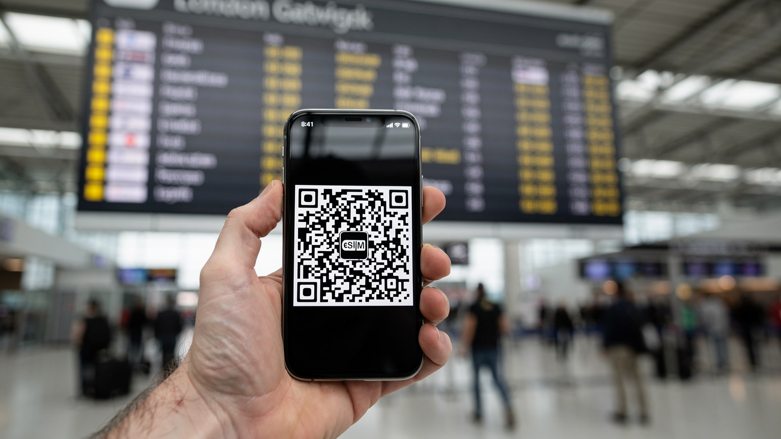 A traveler's hand holding a smartphone showing an eSIM QR code on screen, in front of a departure board at London Gatwick Airport.