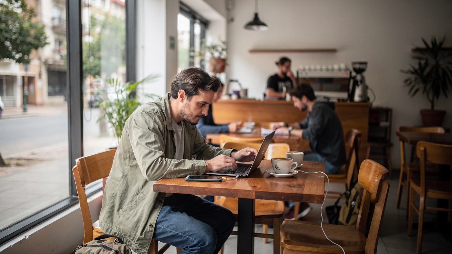 Un viajero trabajando en un café moderno de Montevideo, con su portátil y una taza de café, mientras su móvil se carga discretamente en una mesa de madera.